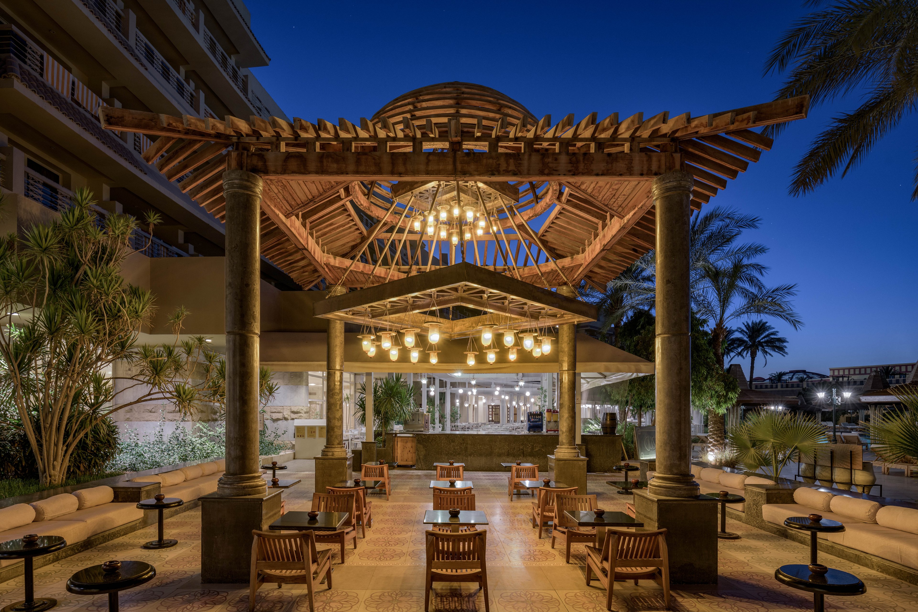 An elegant outdoor dining area featuring a wooden pergola illuminated by warm hanging lights. The setting includes neatly arranged wooden chairs and tables, surrounded by lush greenery and palm trees. The scene is captured during twilight, creating a serene and inviting atmosphere.