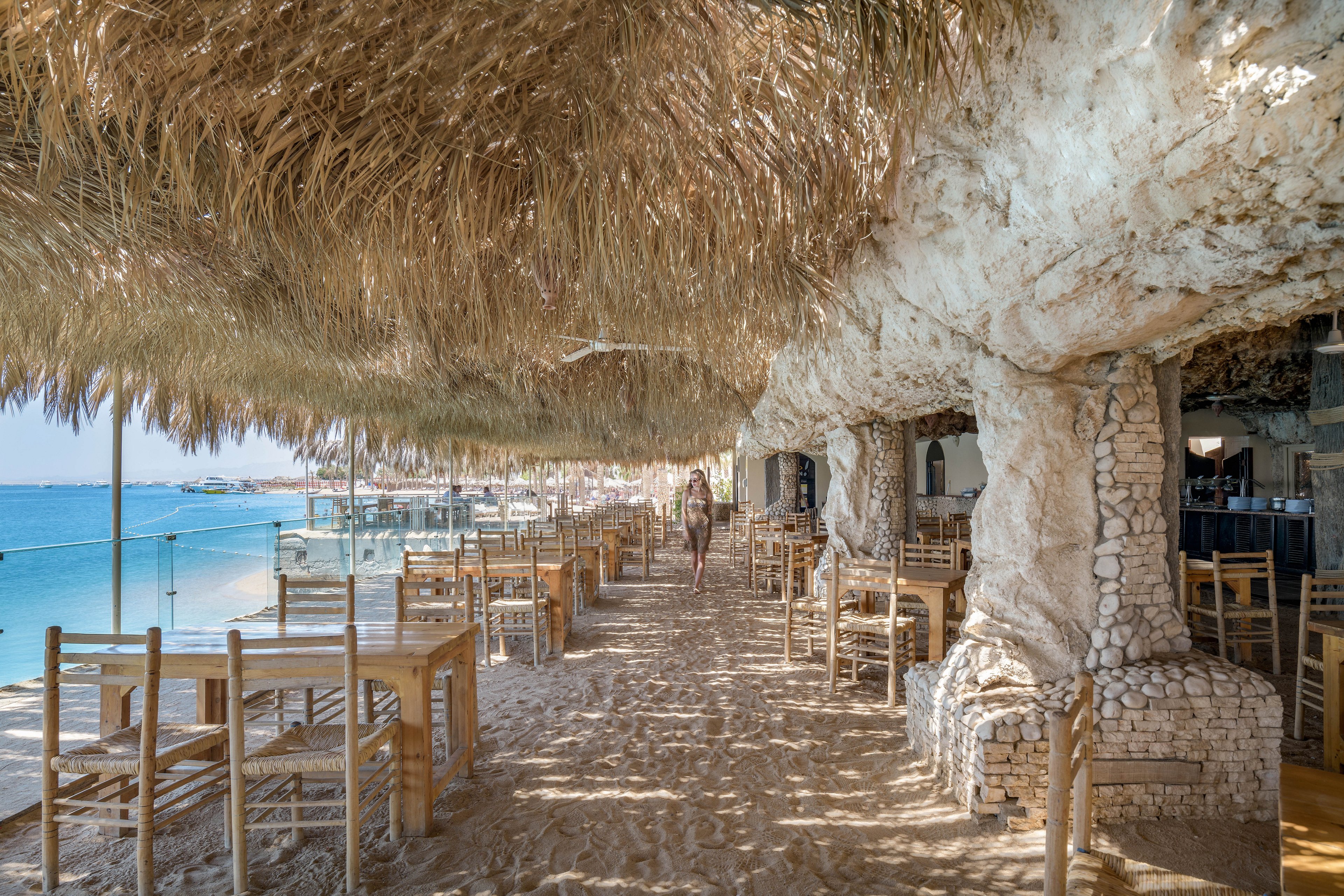 A scenic beachfront restaurant featuring rustic wooden furniture and thatched roofing. The sandy floor and open-air design create a relaxed, tropical atmosphere. The view extends to a calm blue sea, enhancing the serene setting. A single person is visible walking in the distance, adding a sense of scale and activity.
