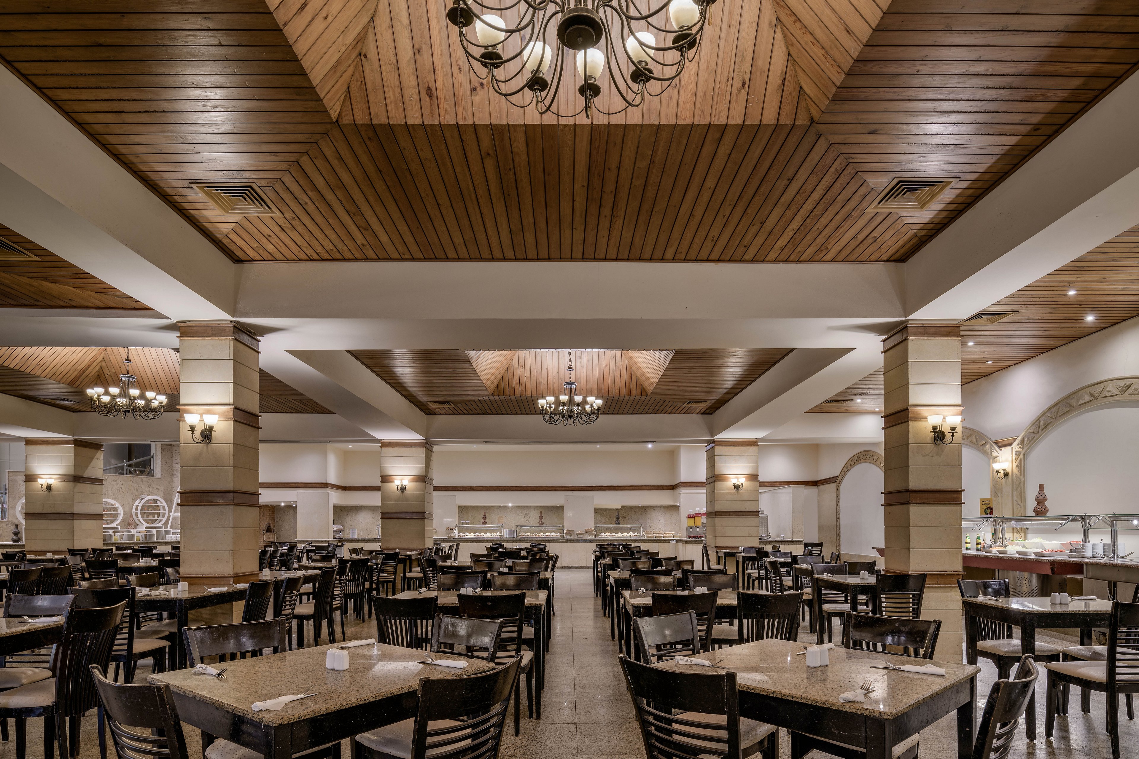 A large, well-lit restaurant dining area featuring a wooden ceiling with intricate chandeliers. The space is furnished with neatly arranged wooden tables and chairs, creating a warm and inviting atmosphere. The background includes a buffet counter and decorative elements, enhancing the elegant setting.