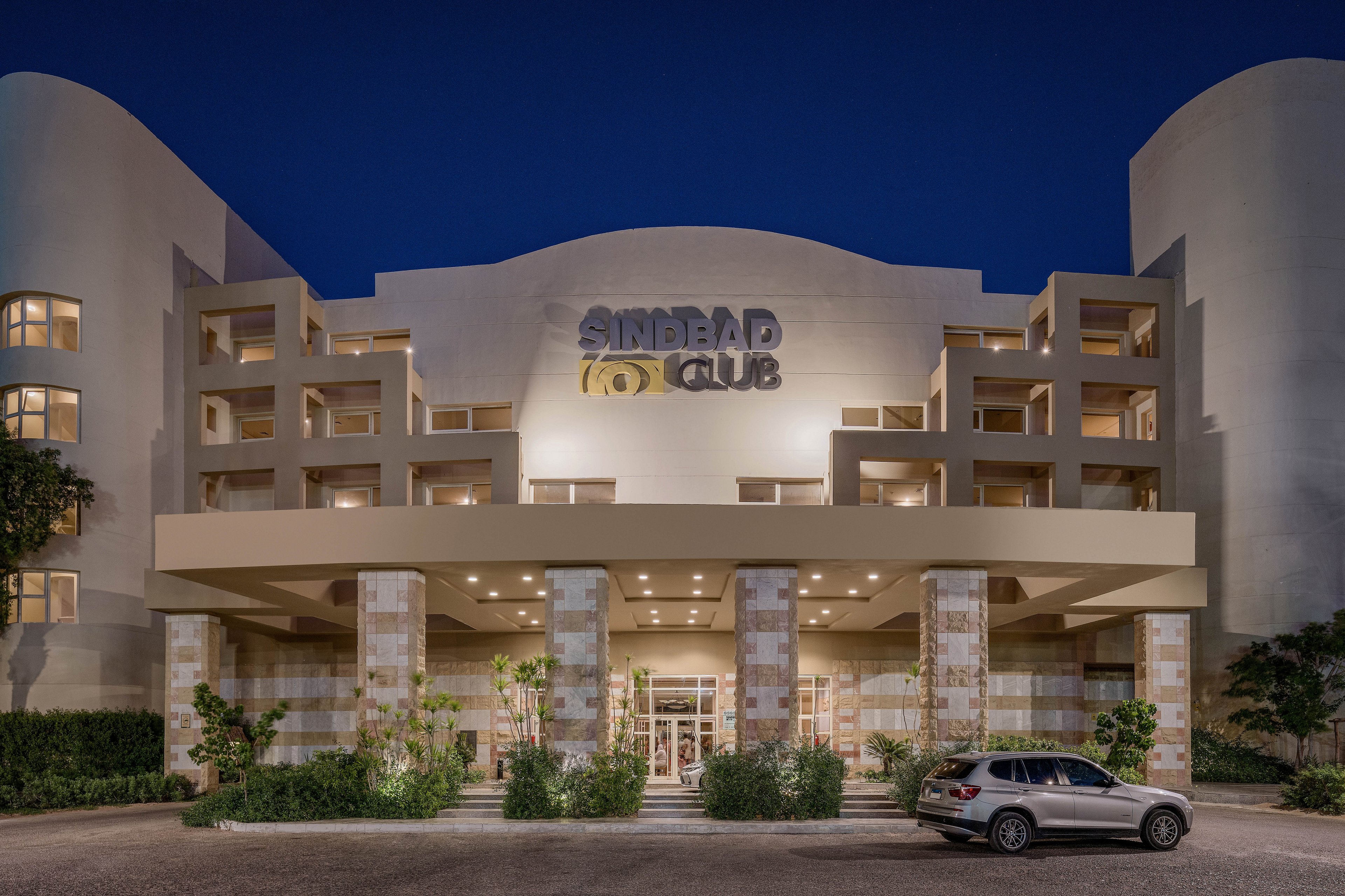A contemporary hotel entrance with a well-lit facade at night, featuring the name 'Sindbad Club' prominently displayed. The building showcases a symmetrical design with balconies and stone columns. A parked car is visible in the foreground, surrounded by greenery.