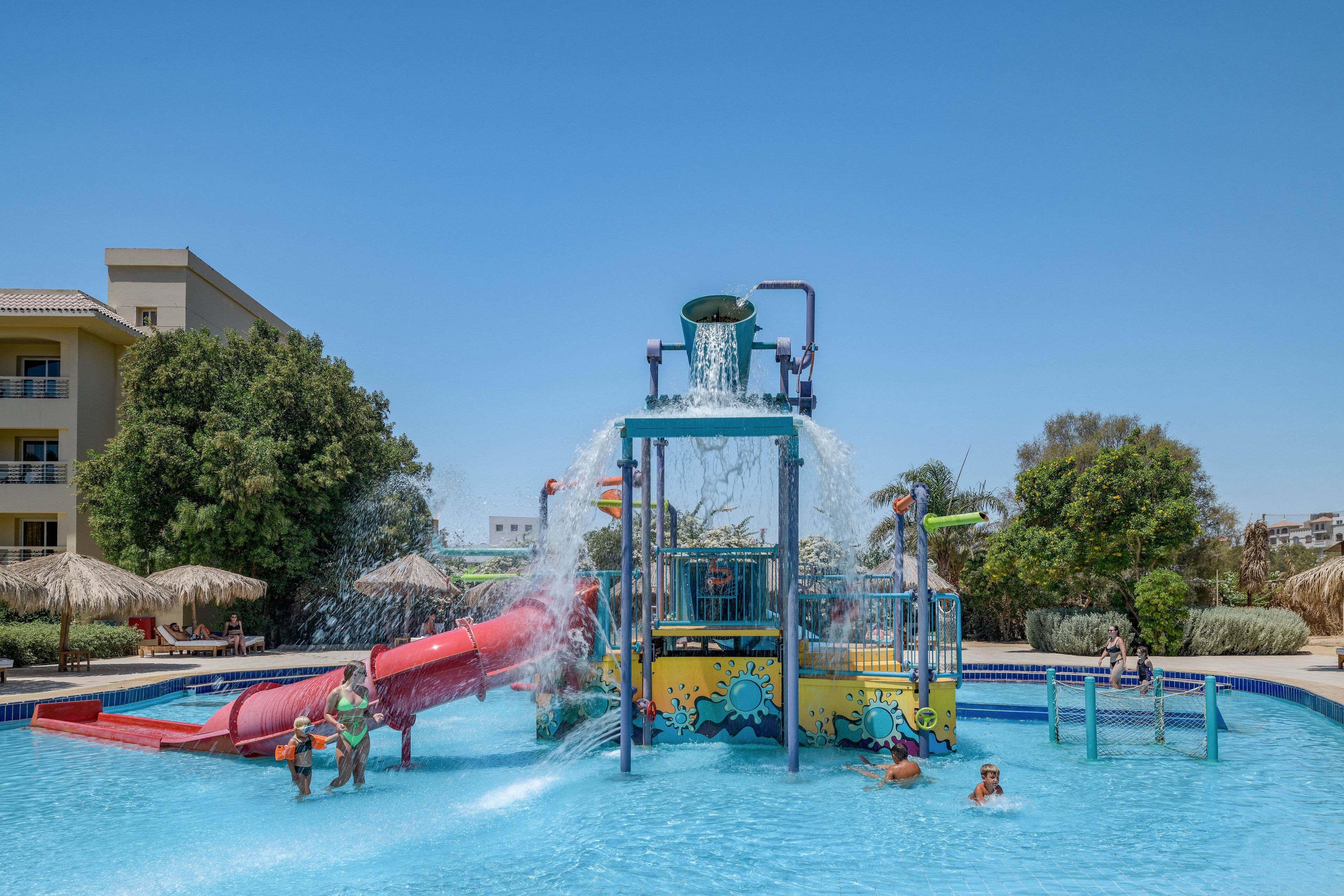 A vibrant water park featuring a central splash tower with cascading water and colorful slides. Children are playing in the shallow pool under a sunny blue sky. The setting includes tropical landscaping and nearby resort buildings, creating a fun and family-friendly atmosphere.