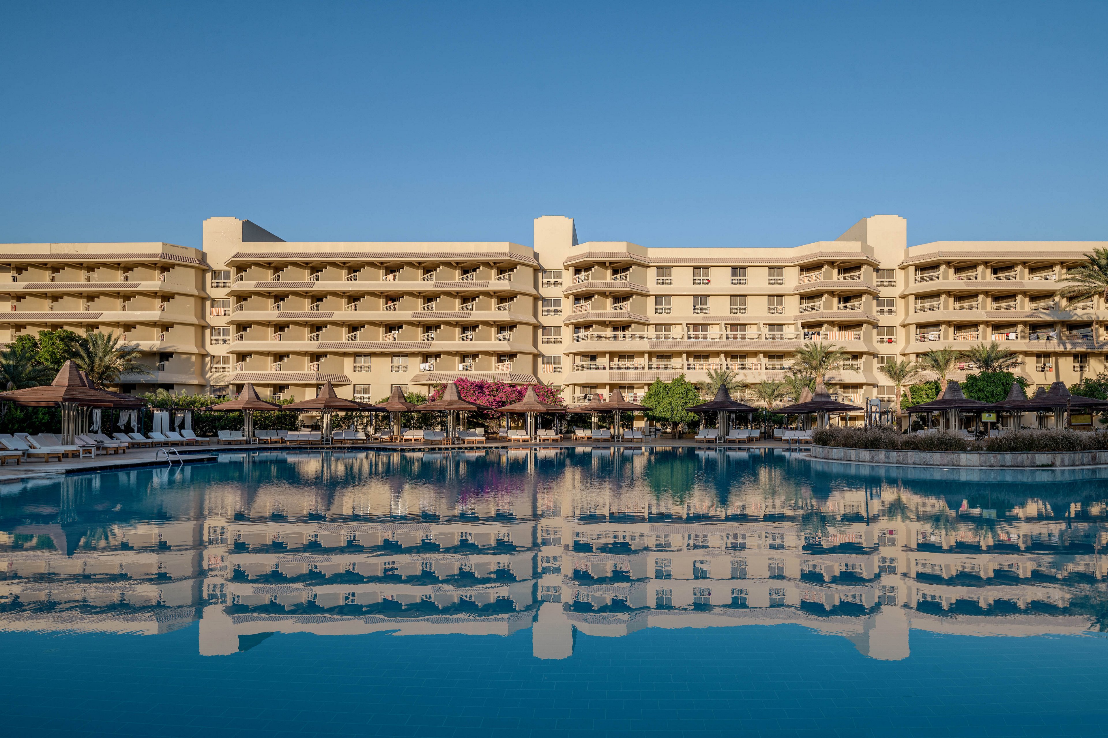 A large luxury resort building is reflected in a calm, expansive swimming pool under a clear blue sky. The setting features lounge chairs, umbrellas, and lush greenery, creating a serene and inviting atmosphere. The symmetrical architecture and tranquil water enhance the visual appeal.