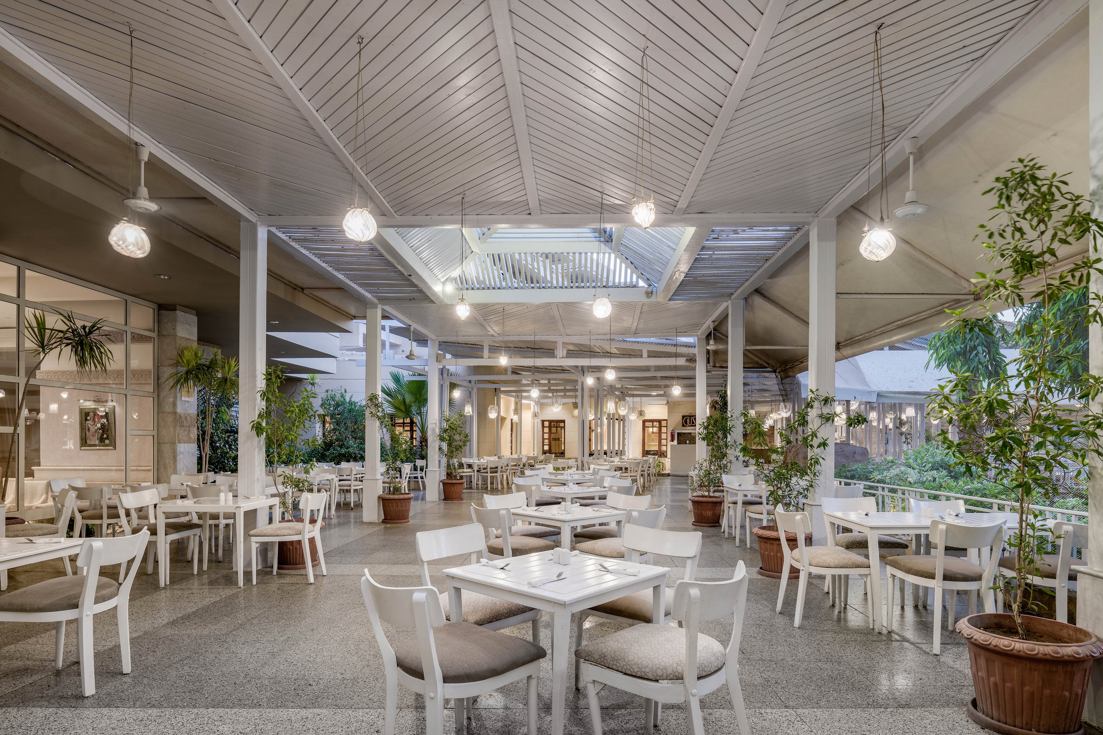 A well-lit outdoor dining area featuring white furniture and a modern design. The space is adorned with hanging lights, potted plants, and a slatted ceiling. The open layout creates a welcoming and airy atmosphere, ideal for casual dining or events.