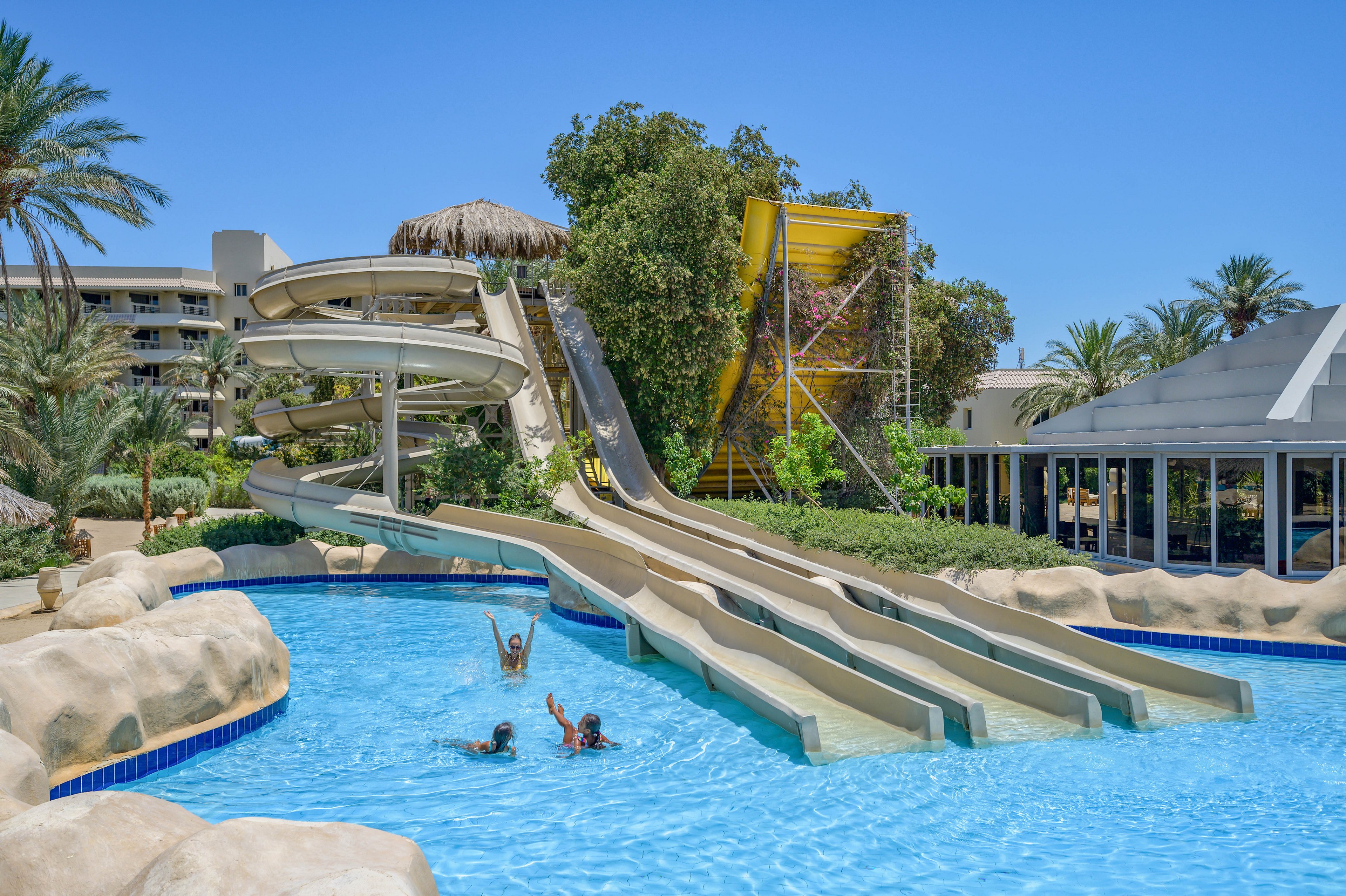 An outdoor water park featuring multiple winding water slides leading into a clear blue pool. Three children are seen enjoying the pool under a sunny sky, surrounded by lush greenery and palm trees. The setting includes a modern building and a vibrant, tropical atmosphere.