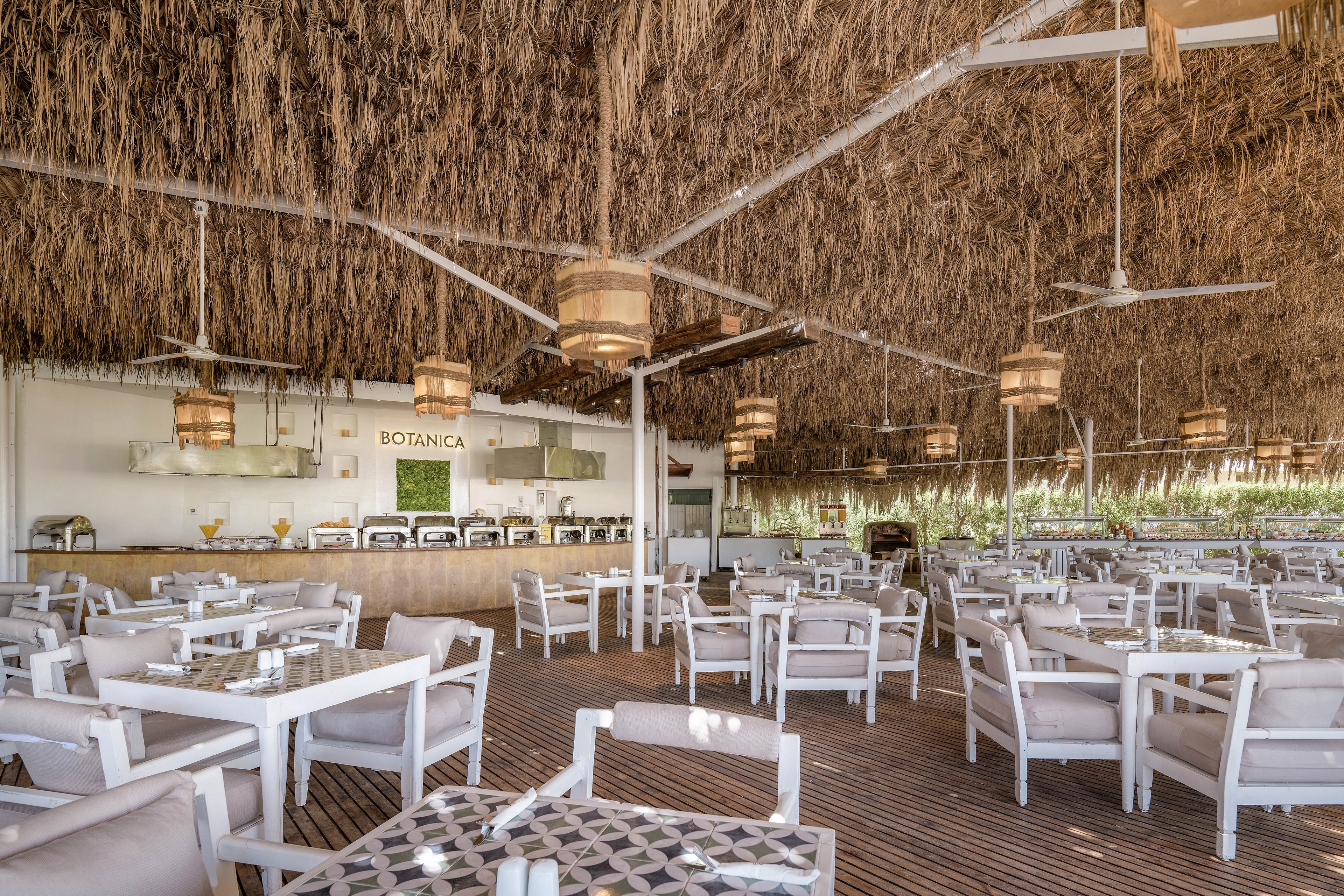 A spacious open-air restaurant featuring a thatched roof and wooden flooring. The seating area includes white tables and cushioned chairs arranged neatly. Warm lighting from hanging lamps creates a cozy atmosphere, and the word 'BOTANICA' is visible on the back wall.