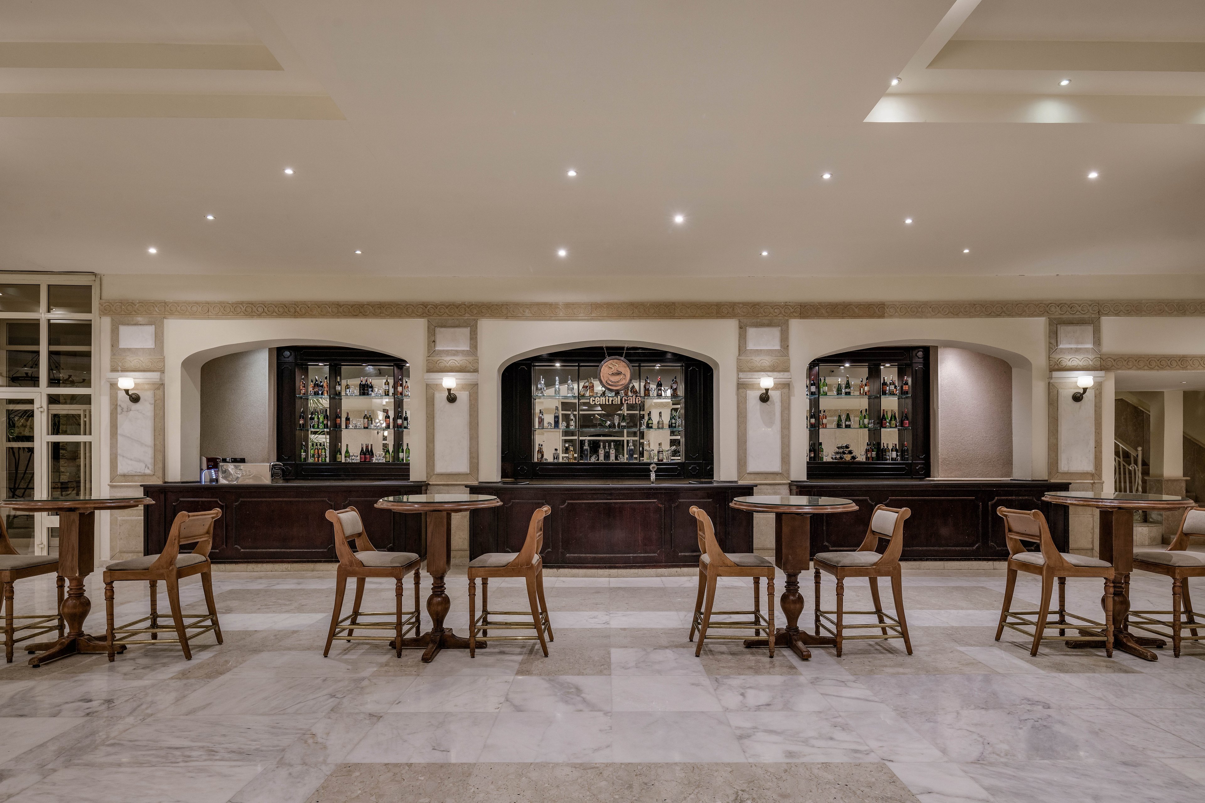 A spacious and well-lit bar interior featuring a long counter with shelves of bottles in the background. Wooden chairs and round tables are neatly arranged on a polished marble floor. The setting exudes a sophisticated and inviting atmosphere, ideal for social gatherings or relaxation.