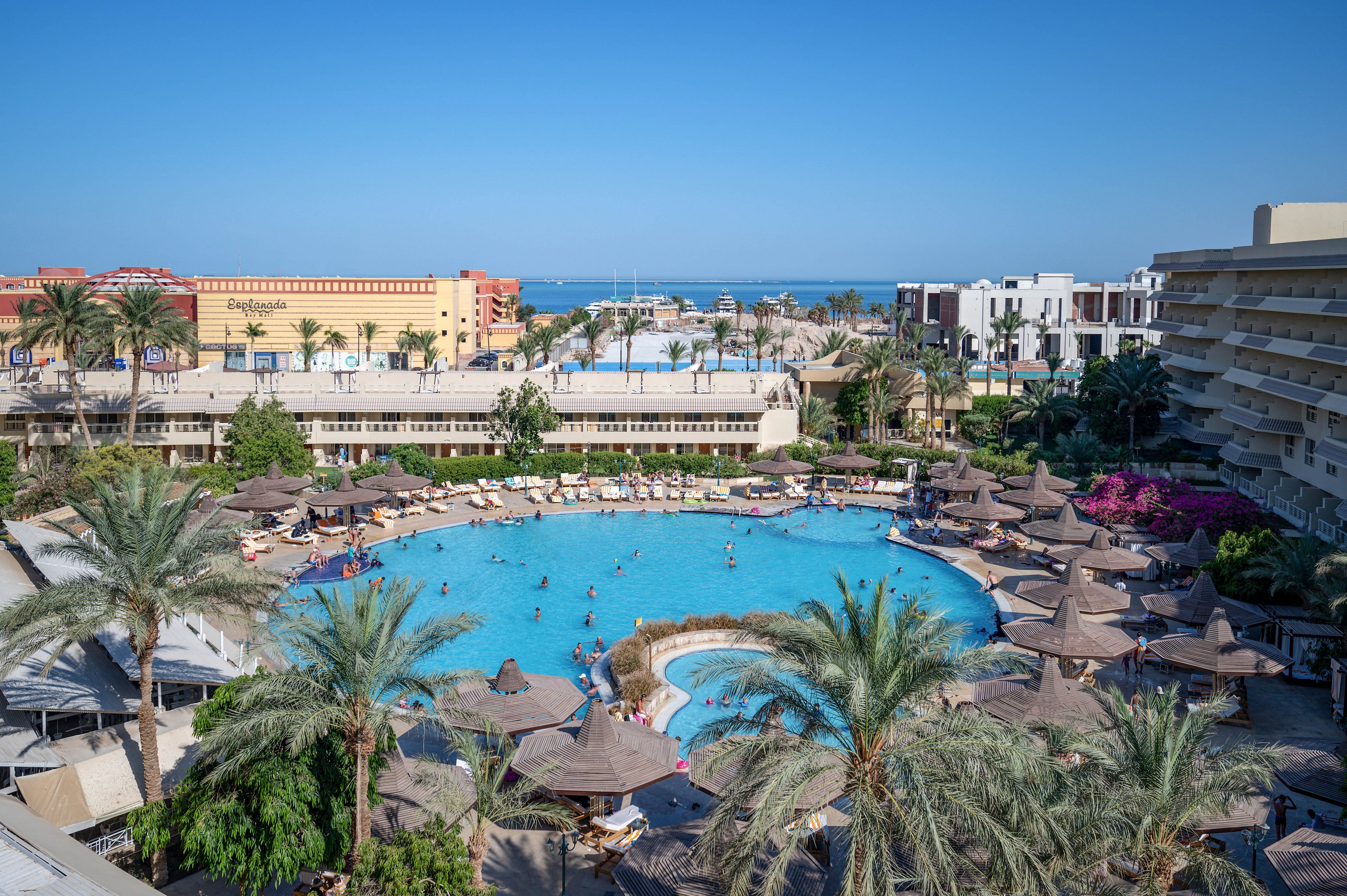 A large outdoor swimming pool surrounded by palm trees and sun loungers in a resort setting. The scene includes multiple buildings, a clear blue sky, and people enjoying the pool. The vibrant atmosphere suggests a tropical vacation destination.