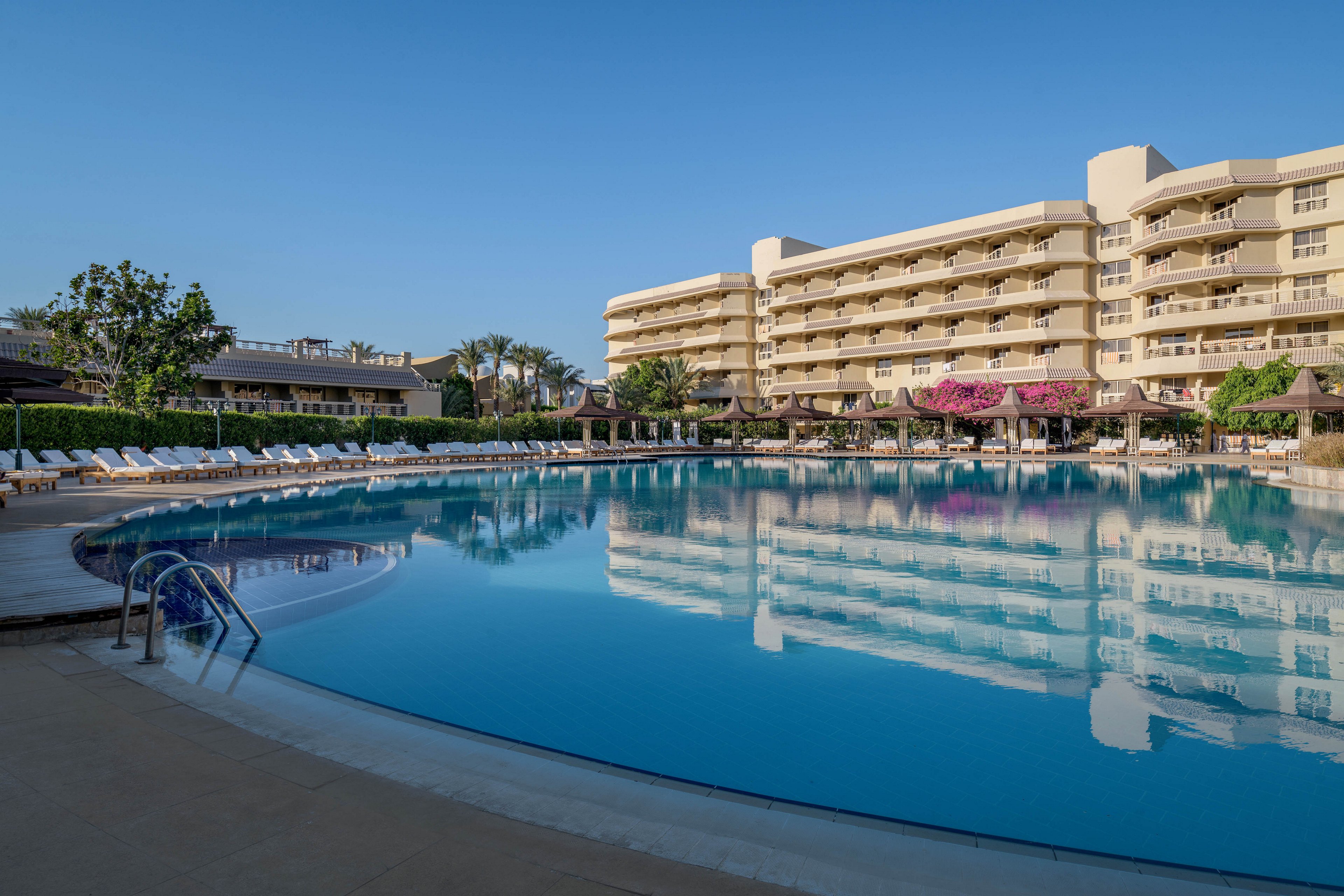 A serene outdoor pool surrounded by lounge chairs and a modern multi-story hotel building. The setting features clear blue skies, lush greenery, and vibrant flowers, creating a relaxing atmosphere. The pool reflects the hotel and surrounding scenery, enhancing the tranquil vibe.