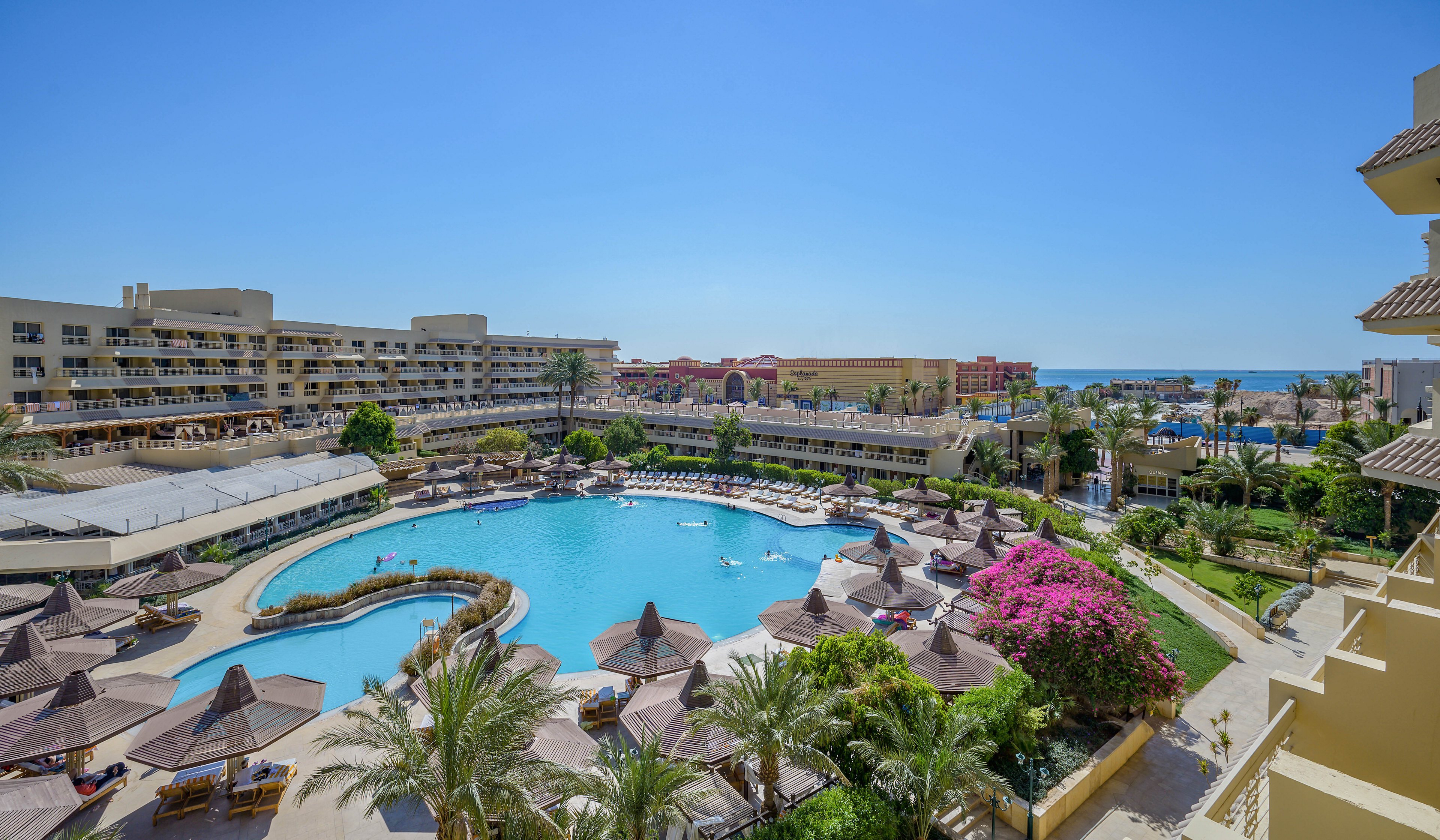 A scenic view of a luxury resort featuring a large, circular swimming pool surrounded by sun loungers and umbrellas. The setting includes lush palm trees, vibrant flowers, and multi-story buildings in the background. The clear blue sky and distant sea add to the tranquil atmosphere.