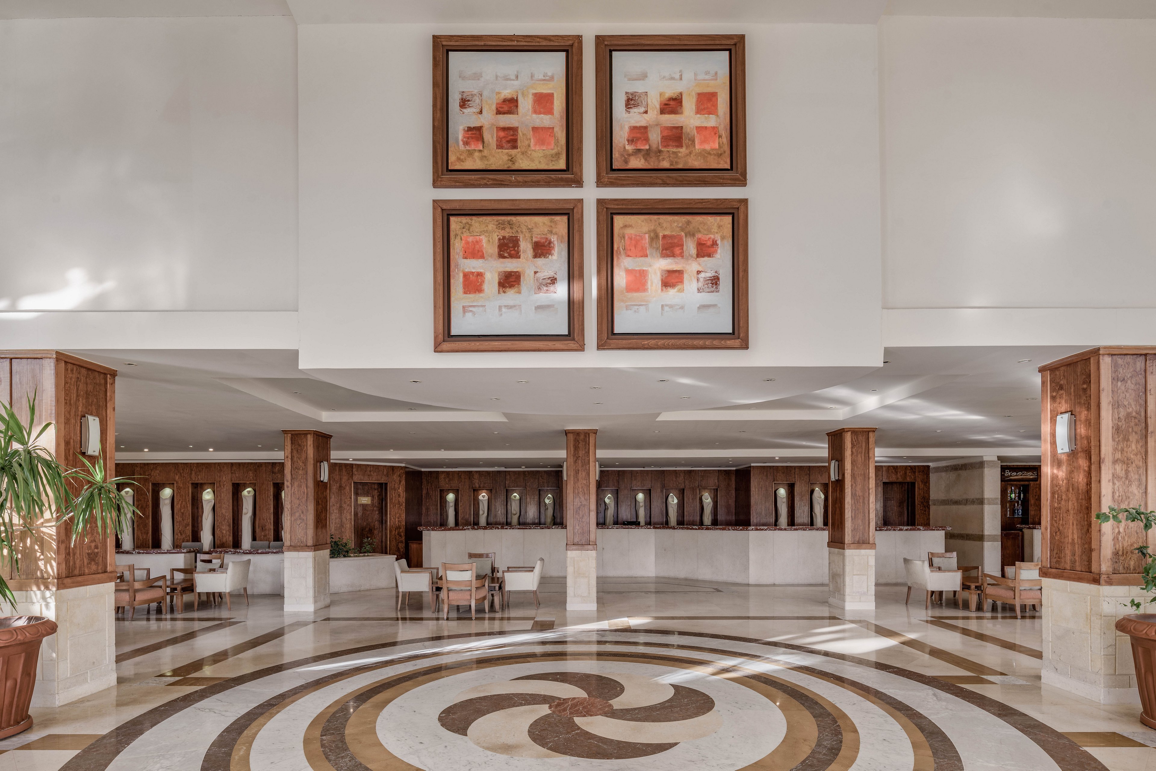 A spacious hotel lobby featuring a circular patterned floor design and modern wooden accents. The walls are adorned with four abstract art pieces, adding a touch of sophistication. The seating area includes neatly arranged chairs and tables, creating a welcoming atmosphere. Natural light enhances the warm tones of the interior.
