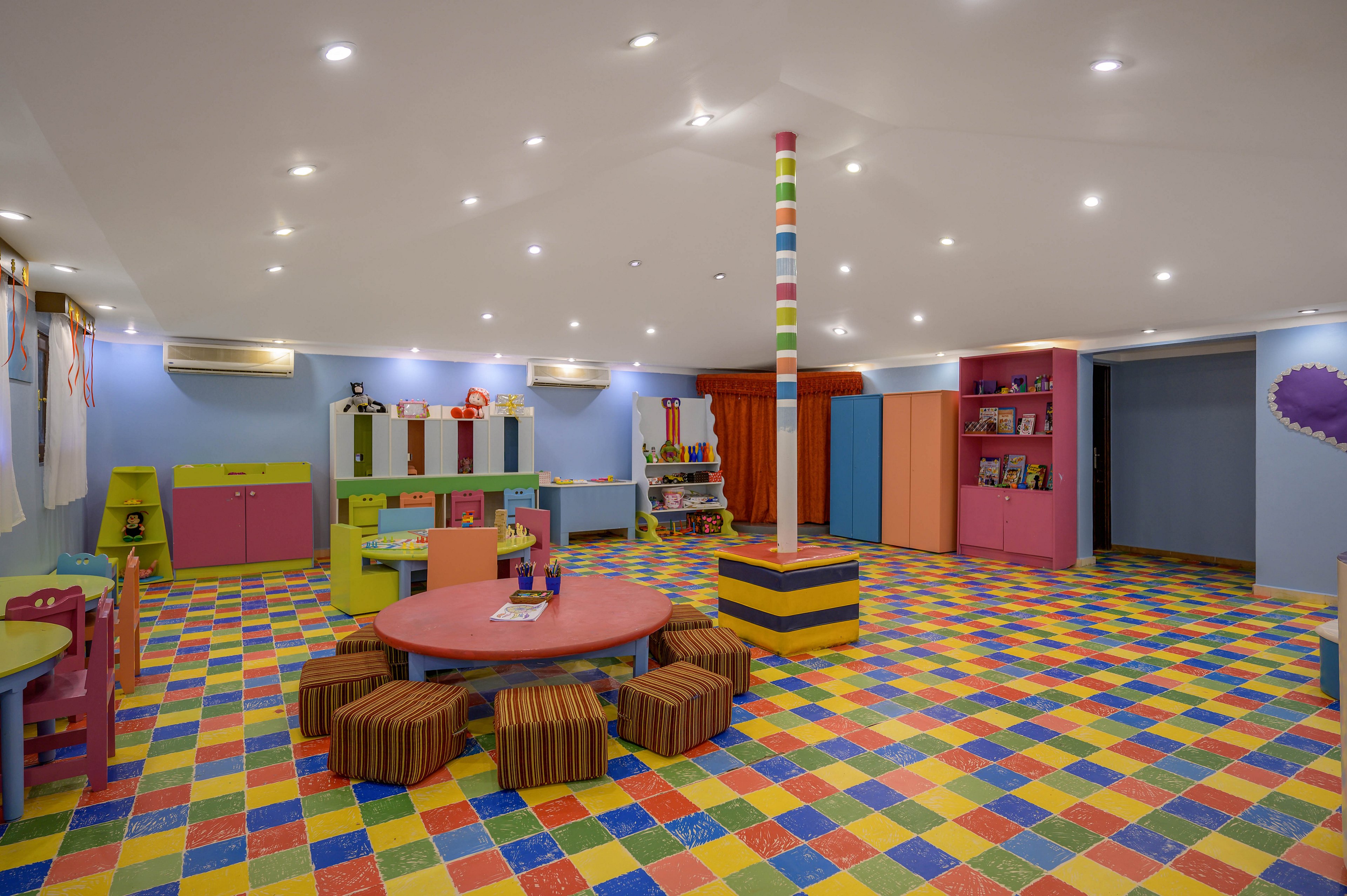 A brightly lit children's playroom featuring colorful furniture and a vibrant tiled floor. The room includes round tables, striped stools, and shelves filled with toys and books. The playful design is enhanced by a multicolored pole and cheerful decor, creating a lively and engaging atmosphere.