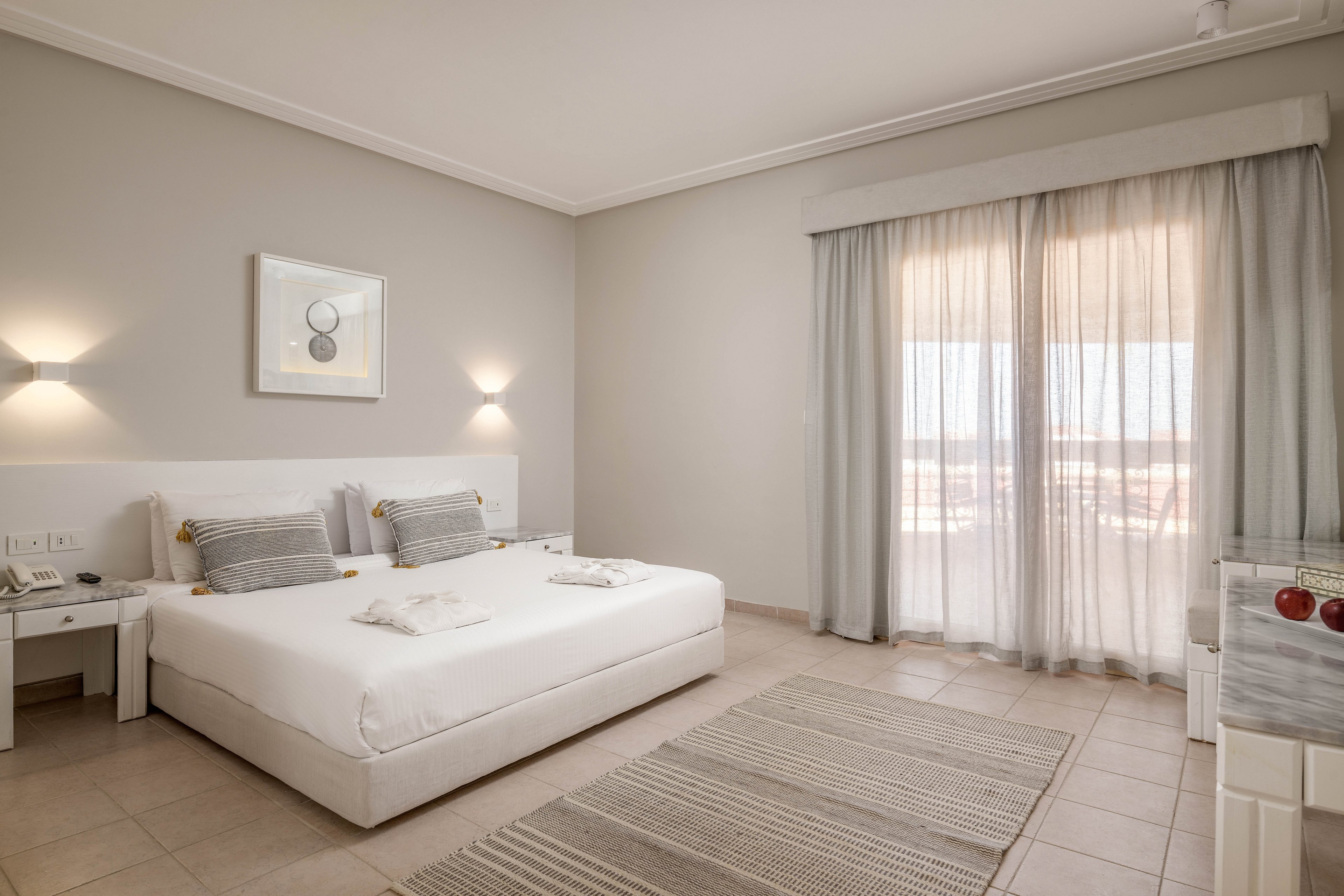 A spacious and modern hotel room featuring a neatly made double bed with gray and white accents. The room is illuminated by natural light streaming through sheer curtains covering a large glass door. Minimalist decor includes wall-mounted lights, a framed artwork, and a neutral-toned rug on tiled flooring.
