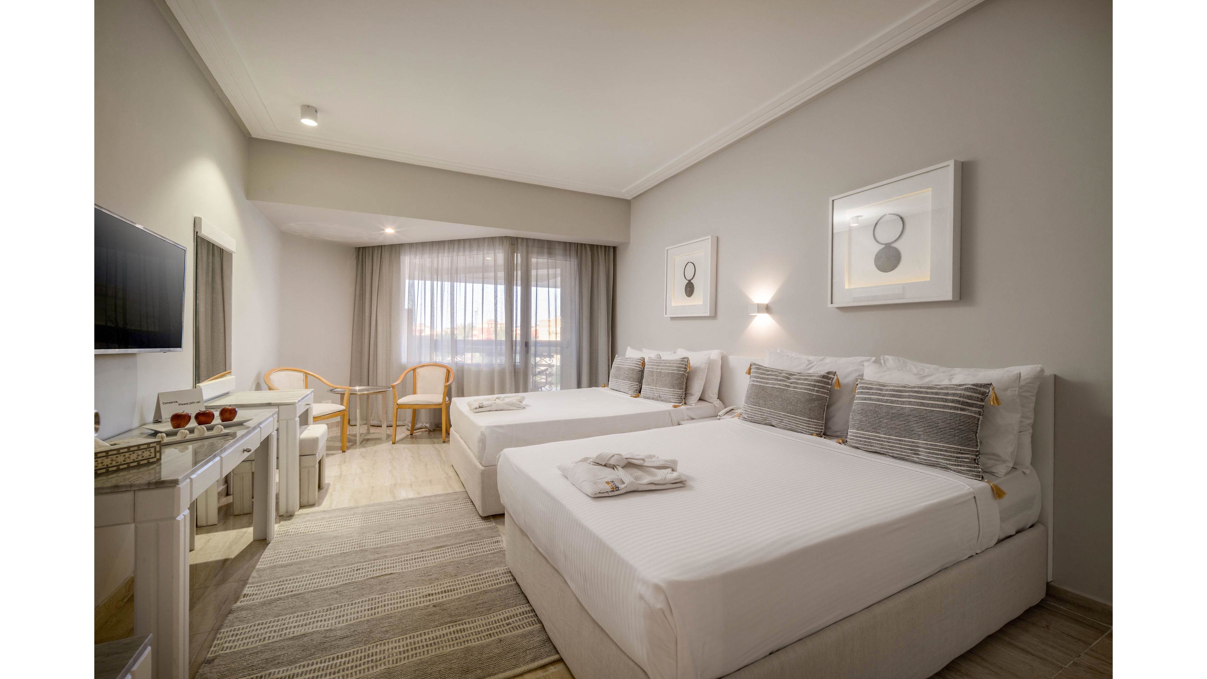 Bright contemporary hotel room featuring two neatly made double beds with gray accent pillows. A large window with sheer curtains lets in natural light and offers a view outside. Minimalist artwork decorates the walls, and a small seating area with wooden chairs sits near the window. A desk with a mirror and television completes the clean, neutral-toned interior.