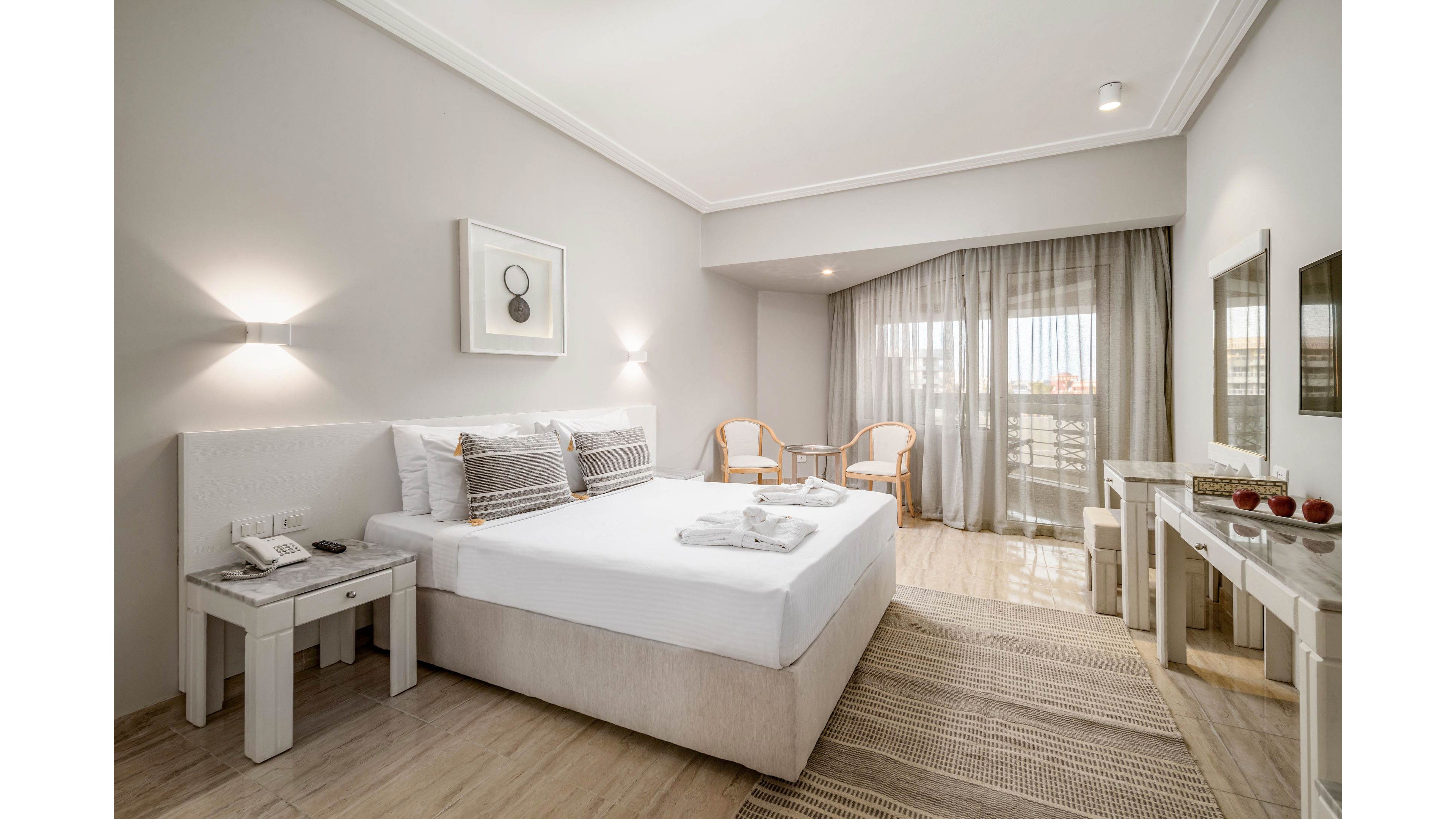 Contemporary hotel bedroom featuring a large double bed with white linens and gray accent pillows. The room includes bedside tables, a desk with mirror, and two chairs with a small table near the balcony. Soft neutral tones, light wood flooring, and sheer curtains create an airy, minimalist atmosphere. Wall art and subtle lighting add a refined, relaxing touch to the interior.