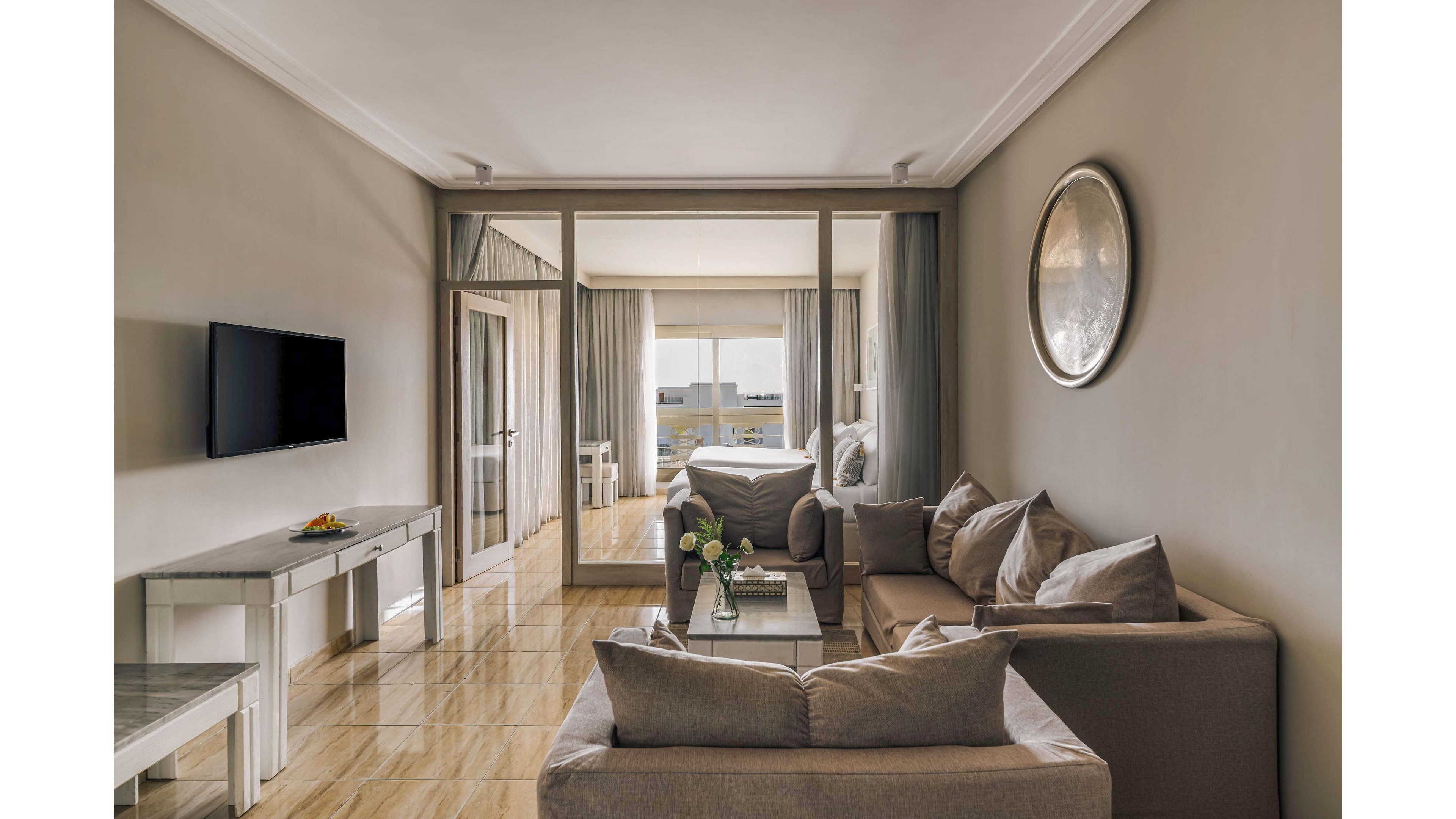 Contemporary hotel living area with beige sofas opens into a bright bedroom through large glass sliding doors. A wall-mounted flat-screen TV faces a sleek console table with a small fruit plate. Soft curtains frame the balcony view, allowing natural light to fill the room. The polished floor and muted neutral palette create a clean, elegant, and relaxing atmosphere.