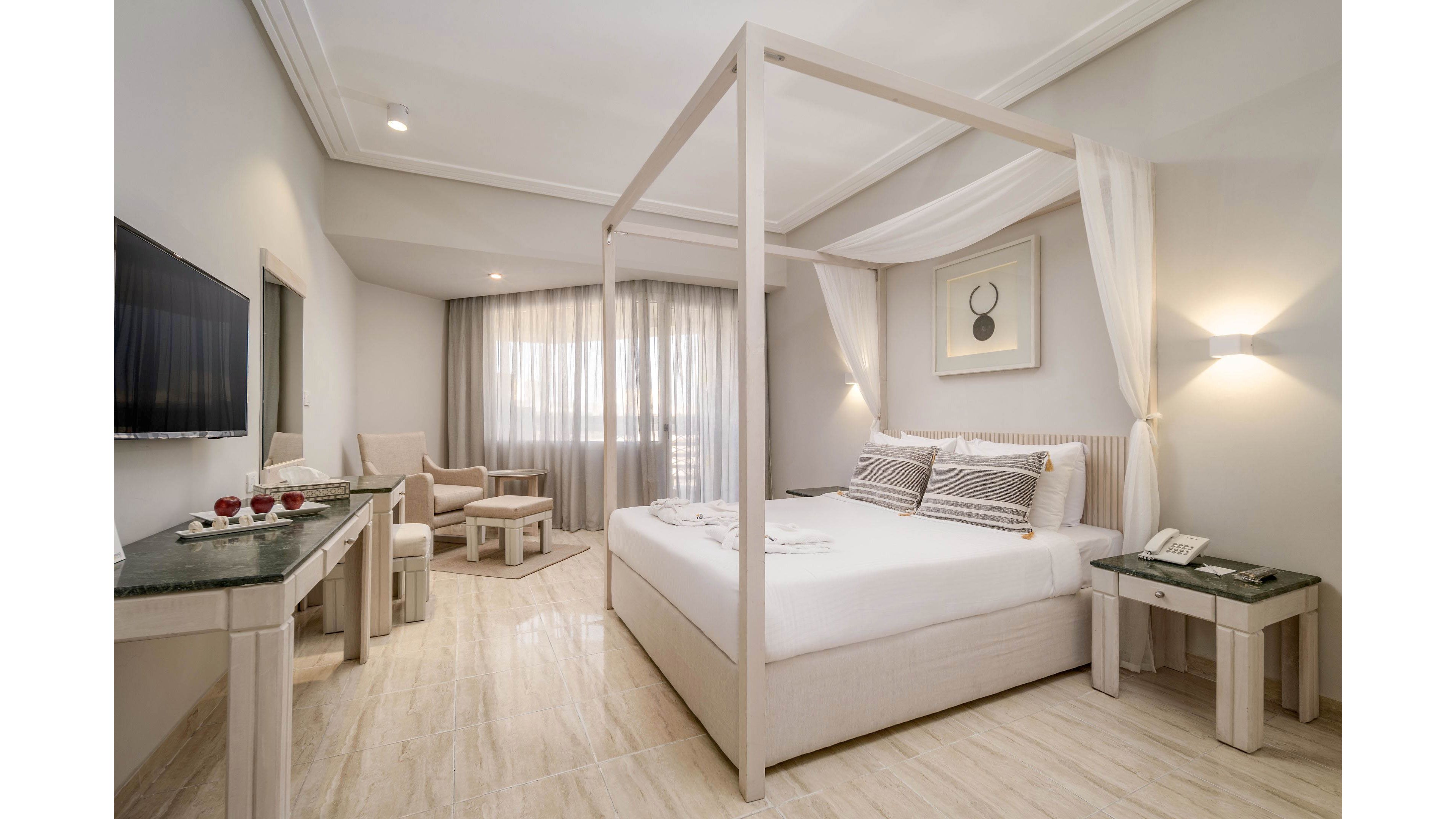 Spacious contemporary bedroom features a white four-poster canopy bed with neatly arranged pillows and linens. The room is decorated in soft neutral tones, with light wood flooring and matching bedside tables and desks. A seating area with armchairs and a small table sits near a large window covered by sheer curtains, allowing natural light to fill the space. Wall-mounted lighting and minimalist artwork add a clean, modern touch to the serene interior.