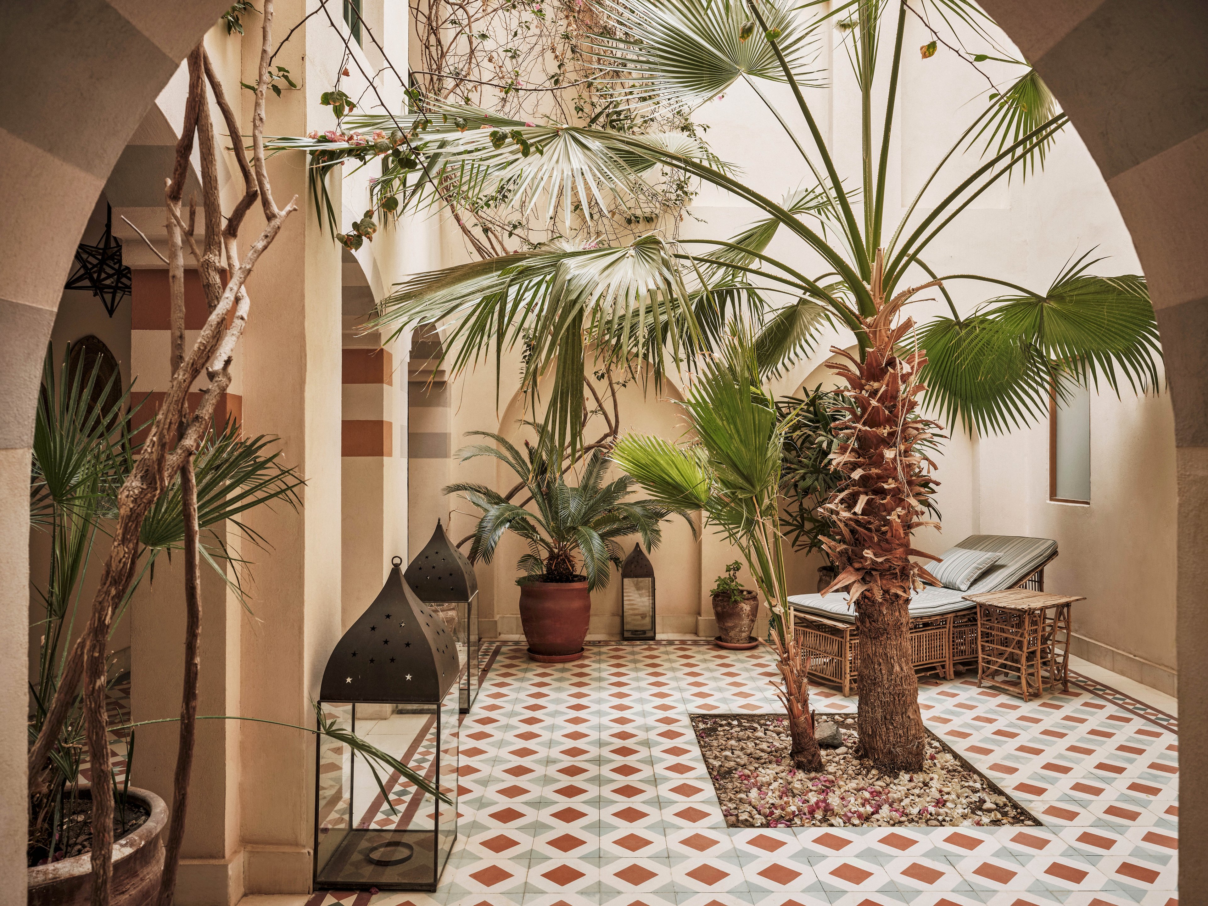 A tranquil courtyard features lush palm trees and various potted plants, creating a relaxing oasis. The space is defined by geometric patterned floor tiles in earthy tones, with natural light filtering through the open ceiling. Wicker lounge chairs and a large decorative lantern add to the inviting, Mediterranean-inspired atmosphere.