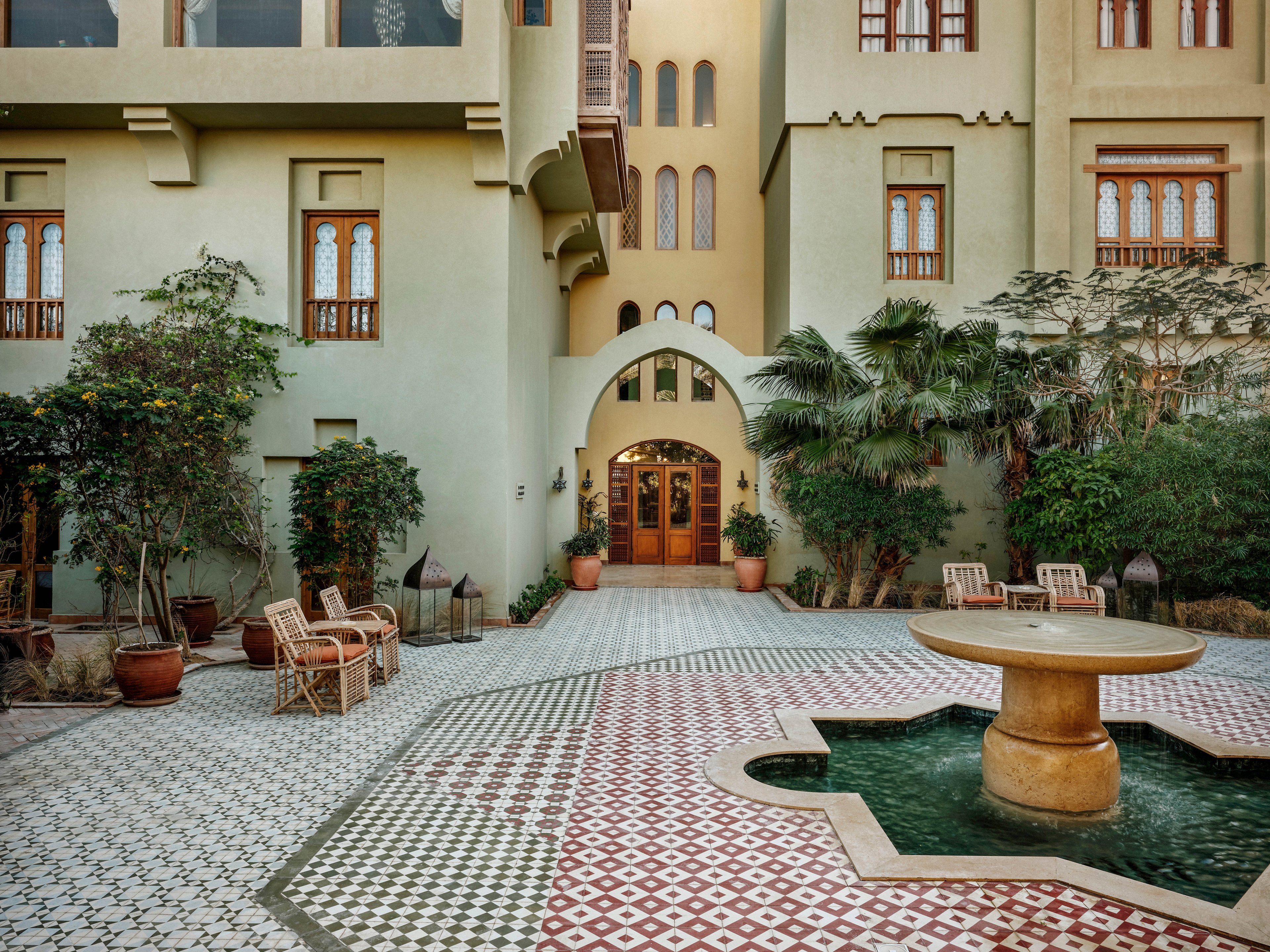 A serene courtyard features a geometric patterned tile floor and a round stone fountain at its center. The setting is surrounded by lush greenery, potted plants, and classic wooden windows on a beige building facade. Several wicker chairs and tables are arranged for relaxation, creating a tranquil and inviting atmosphere. The architectural style suggests a blend of traditional and modern influences.