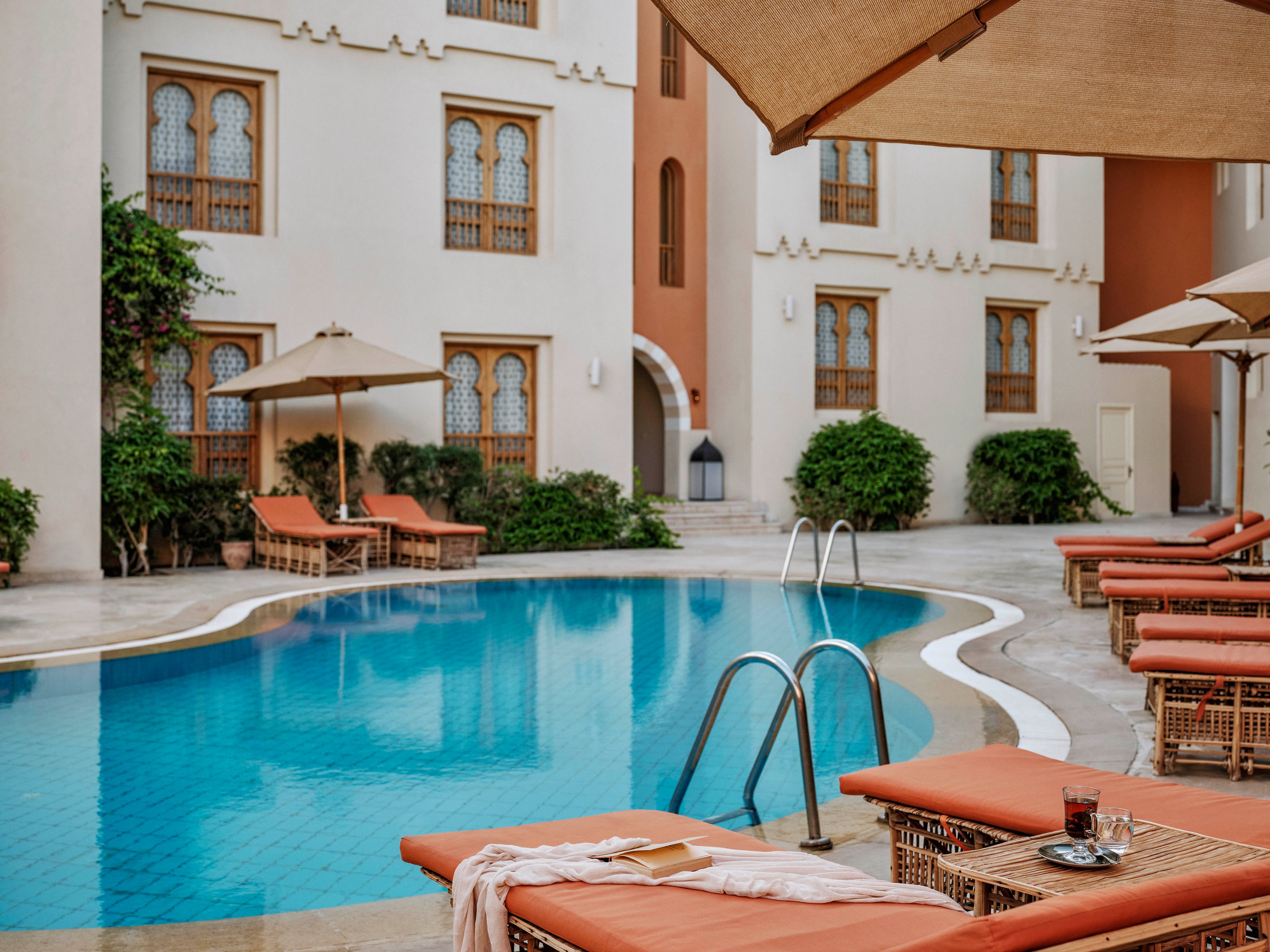 A tranquil outdoor swimming pool is surrounded by cushioned lounge chairs and large umbrellas in a courtyard setting. The architecture features arched windows and decorative details, with lush greenery adding to the relaxing atmosphere. A towel and drinks rest on a table, suggesting leisure and comfort. The scene is calm, with warm earthy tones and soft natural light.