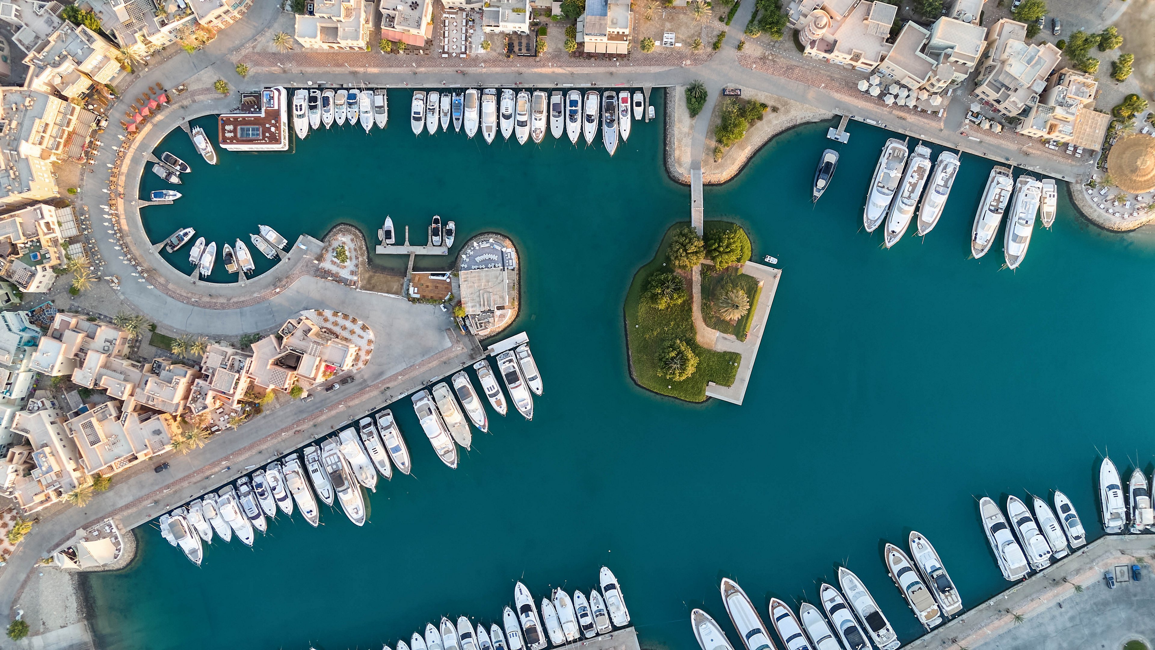 High-angle aerial photograph captures a marina filled with numerous yachts and boats docked along curved piers. The marina is surrounded by waterfront buildings and a small green island at the center, all set in calm blue water. The image highlights the organized layout of the docks and the tranquil atmosphere of the harbor.