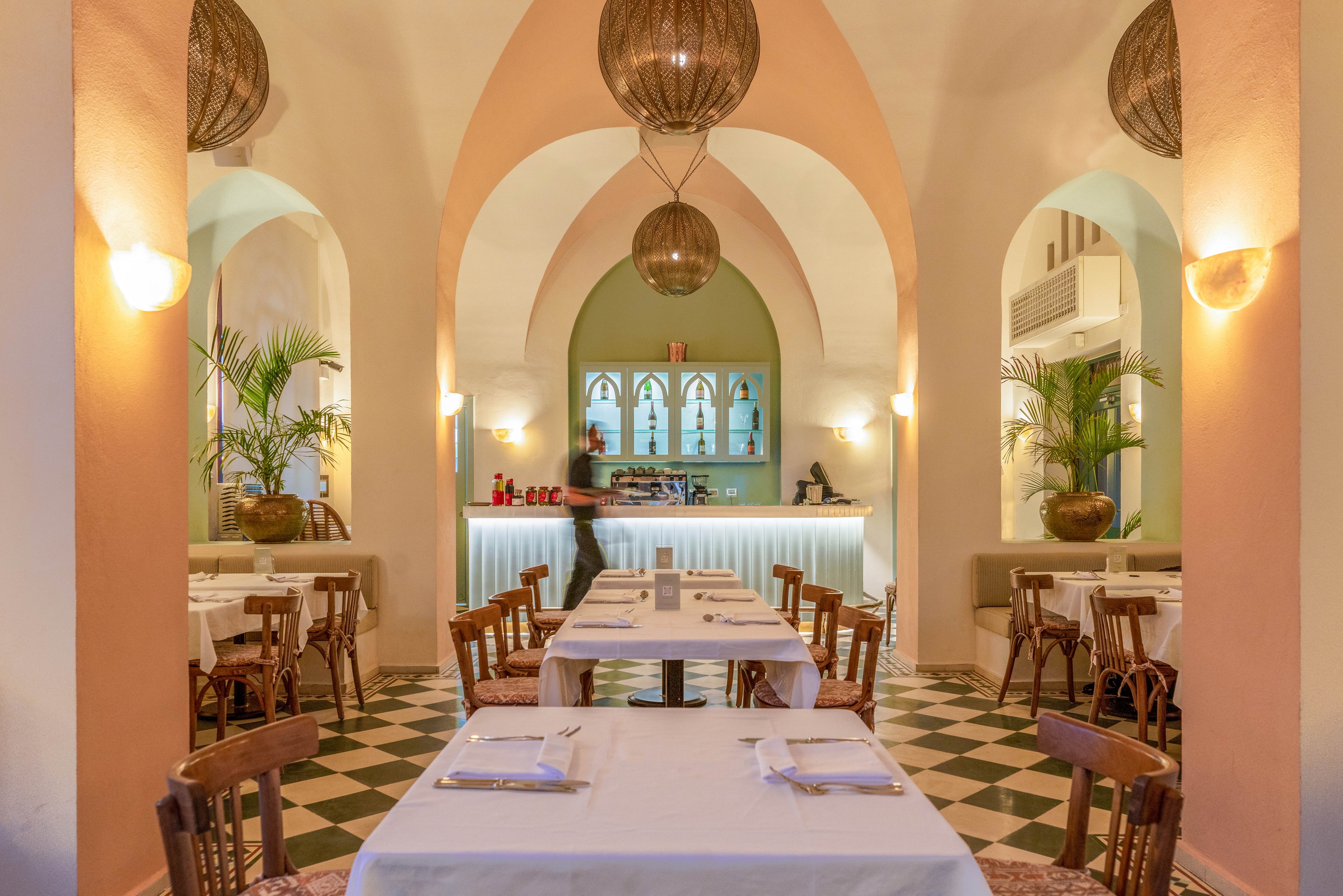 A stylish restaurant dining area features arched ceilings, soft peach and cream walls, and checkered tile flooring. Wooden chairs and tables with white tablecloths are neatly arranged, while large potted plants add a touch of greenery. The softly lit bar at the back creates a welcoming ambiance, enhanced by warm pendant lighting and decorative wall sconces.