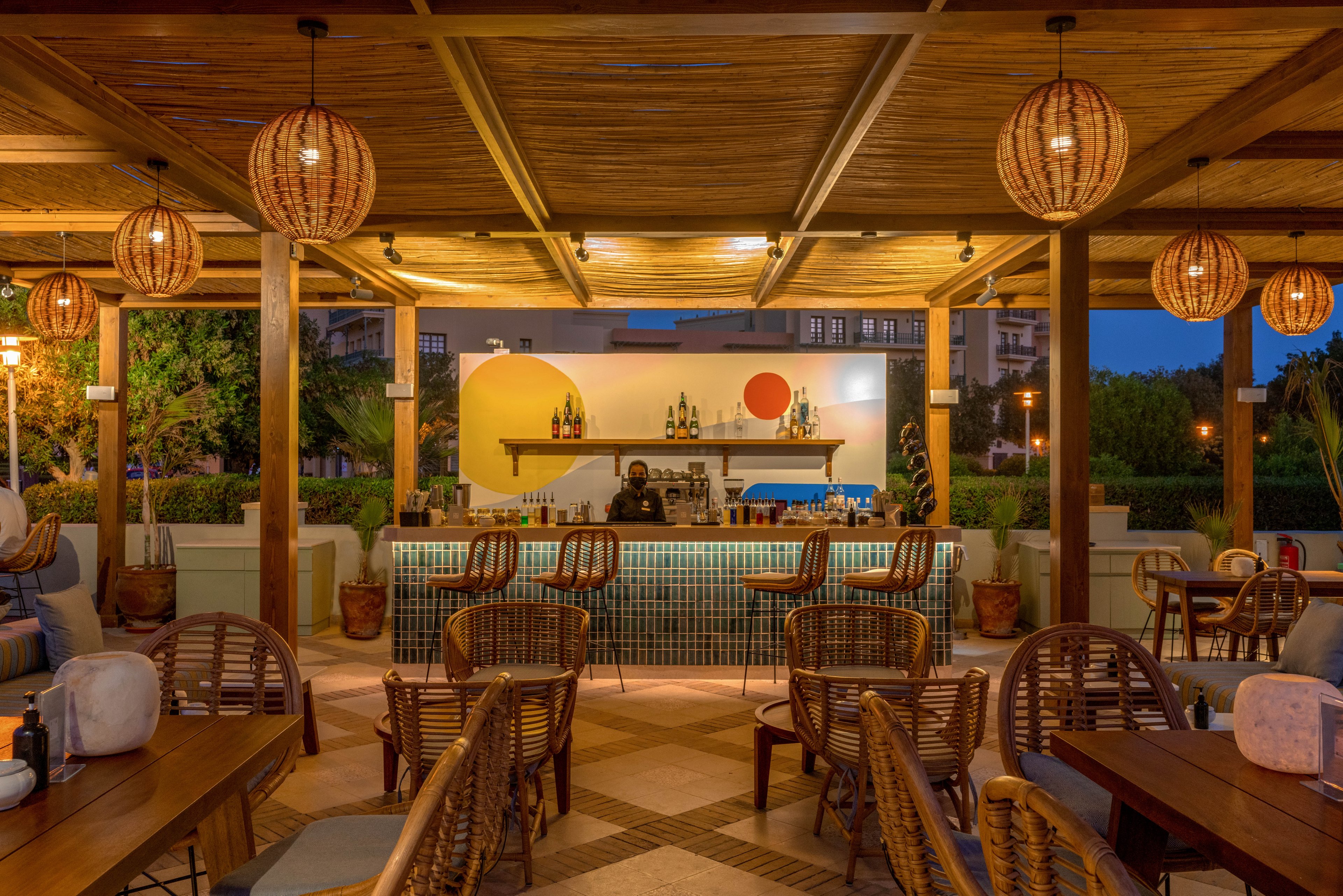 An inviting outdoor bar lounge features woven rattan chairs and wooden tables under a wooden pergola. Warm pendant lights create a cozy atmosphere as evening falls, with a bartender preparing drinks behind a tiled bar. The backdrop includes modern wall art with geometric shapes and bottles displayed on shelves, while lush greenery surrounds the setting.