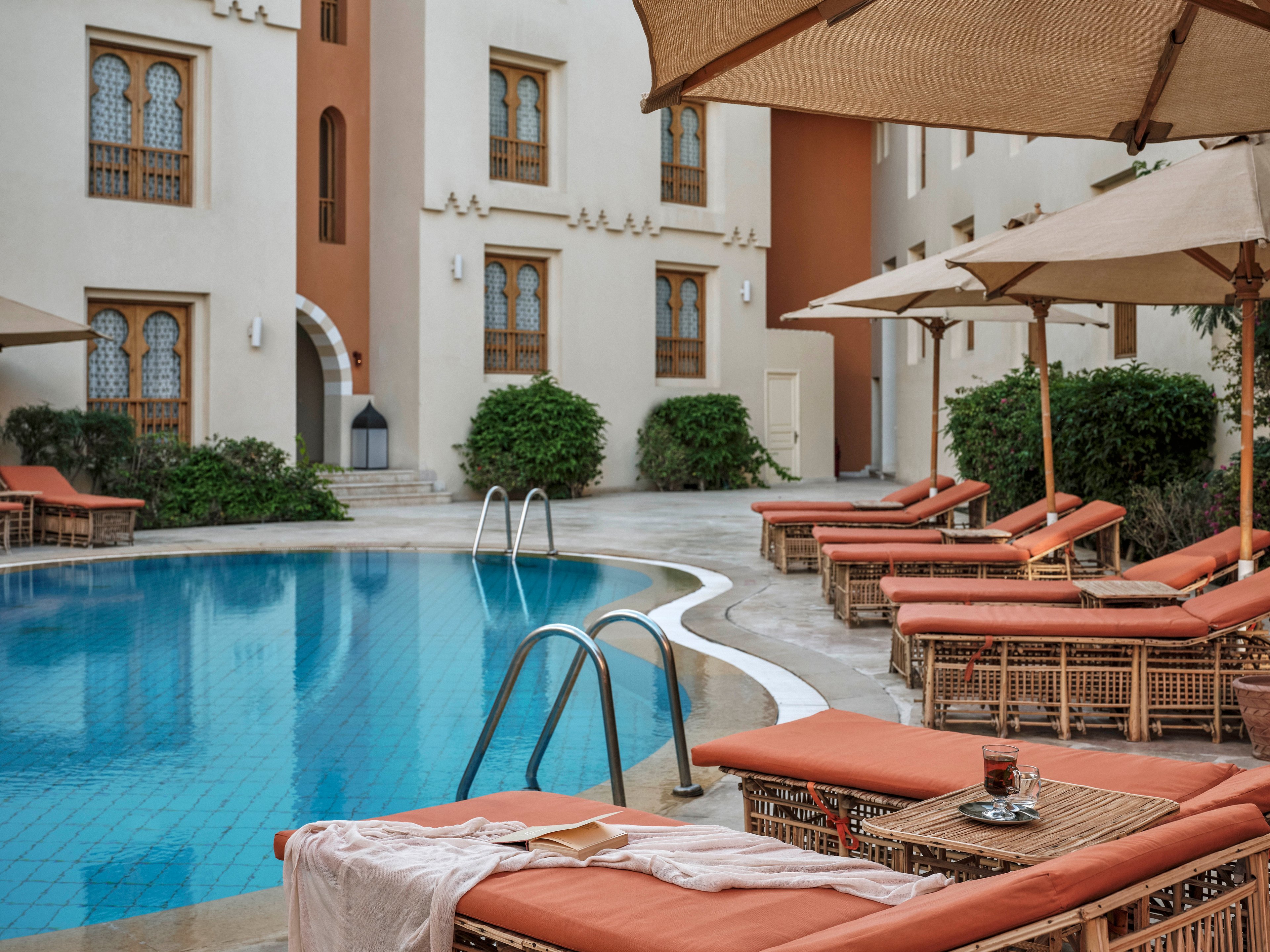 A serene hotel pool area features several cushioned lounge chairs and large umbrellas arranged around a clear blue swimming pool. The setting is tranquil, with Mediterranean-style architecture and lush greenery in the background. A towel and a drink rest on one of the loungers, suggesting relaxation and leisure. The scene is empty of people, emphasizing a peaceful and inviting atmosphere.