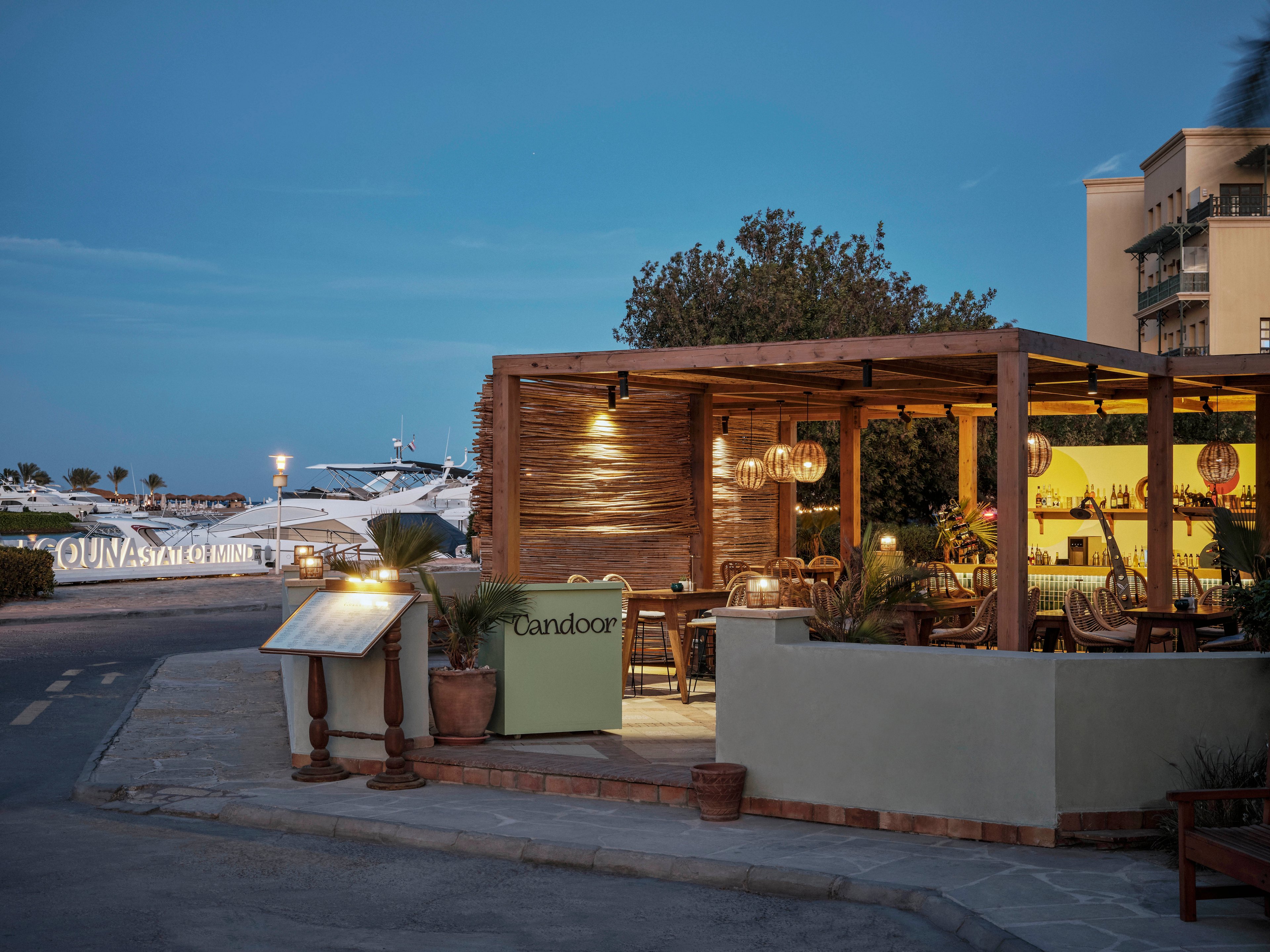 A cozy outdoor restaurant terrace named 'Candoor' is illuminated by warm hanging lights at dusk. The setting is adjacent to a marina with large yachts visible in the background, creating a relaxed and upscale atmosphere. The scene features wooden furniture, potted plants, and a soft blue evening sky. The restaurant sign 'Candoor' is clearly visible at the entrance.