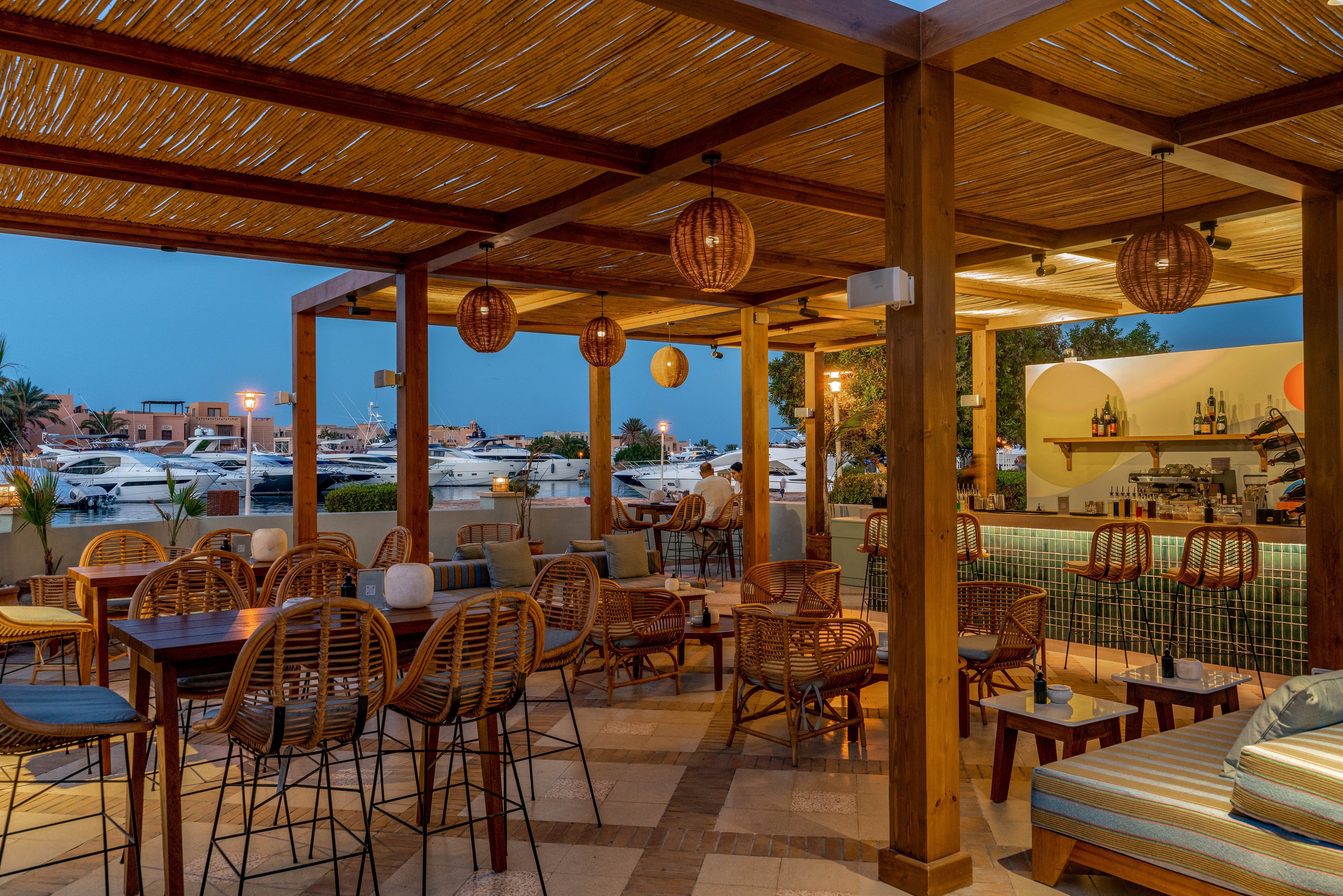 An inviting outdoor bar area features wooden pergola roofing and rattan furniture, set against a marina backdrop with docked boats. Warm pendant lighting creates a cozy ambiance as evening falls. The bar is stocked and ready for guests, with a few people seated and enjoying the relaxed atmosphere. The setting combines natural wood tones with soft blue and beige accents, enhancing the tranquil, upscale vibe.