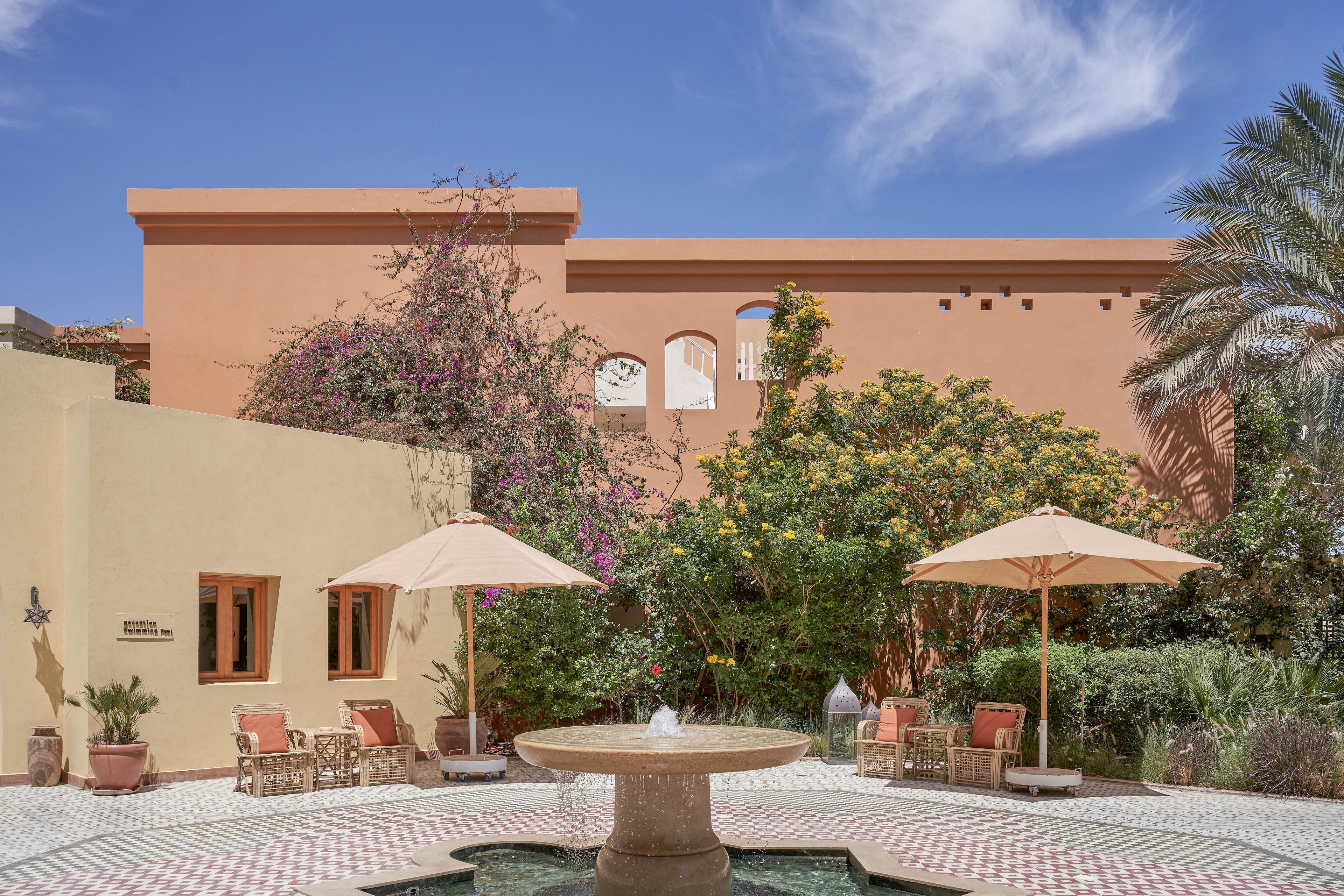 A tranquil courtyard features a central stone fountain surrounded by patterned tile flooring. Two beige umbrellas shade seating areas with chairs and tables, set against lush greenery and flowering plants. The backdrop includes a terracotta-colored building with arched windows, evoking a Mediterranean or North African architectural style. The sky is clear and blue, enhancing the bright, relaxed atmosphere.