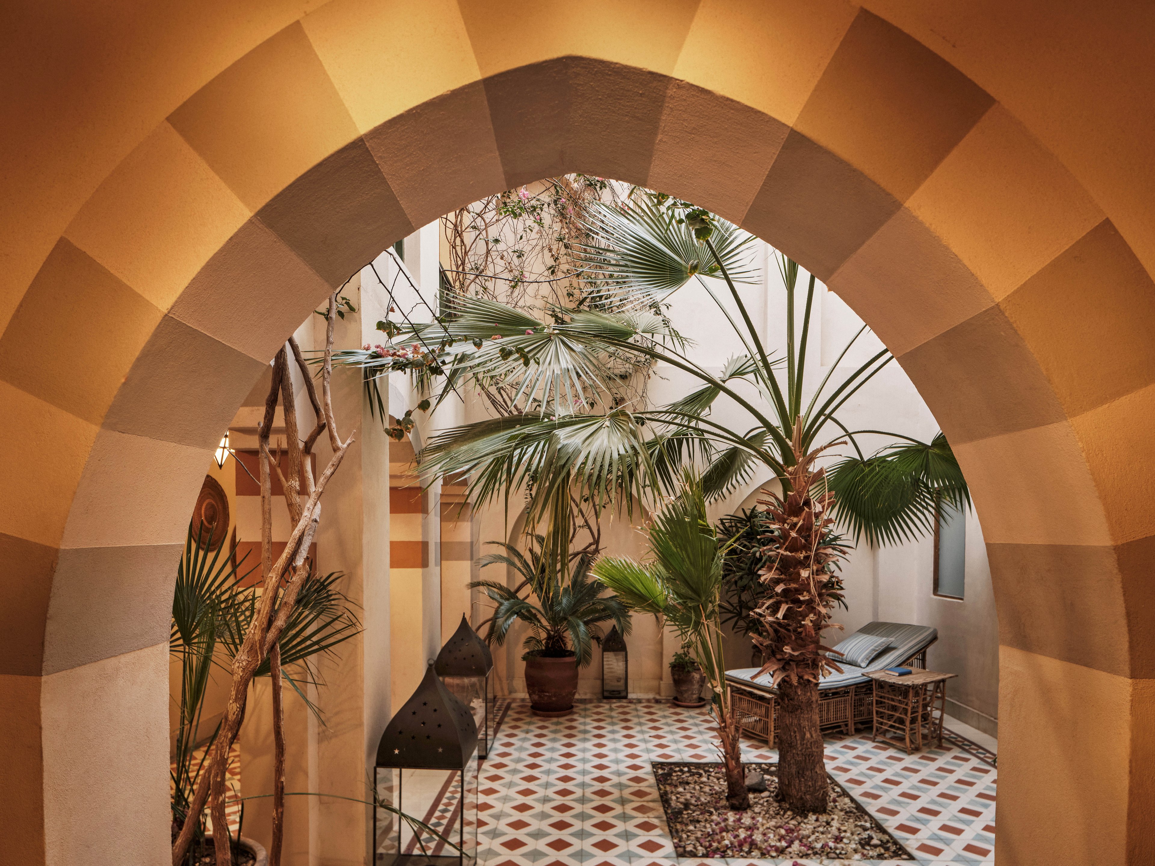 A traditional Moroccan courtyard is viewed through a large arched entryway. The space features patterned tile flooring, several potted palm trees, and a mix of natural and architectural elements. Warm earth tones and geometric designs create a serene, inviting atmosphere. No visible text or numbers are present in the image.
