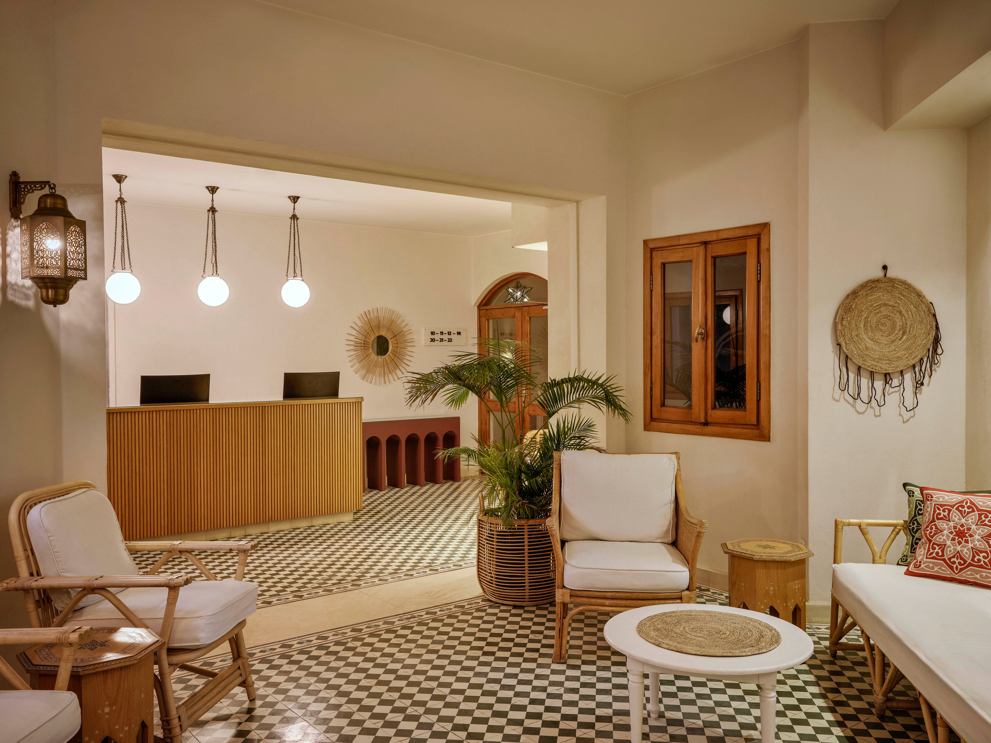 A warmly lit hotel lobby features a checkered black and white tile floor and a wooden reception desk with three pendant lights above. The seating area includes cushioned chairs, a small round table, and decorative wall accents such as woven art and a lantern. Potted plants and natural wood elements contribute to a relaxed, inviting atmosphere.