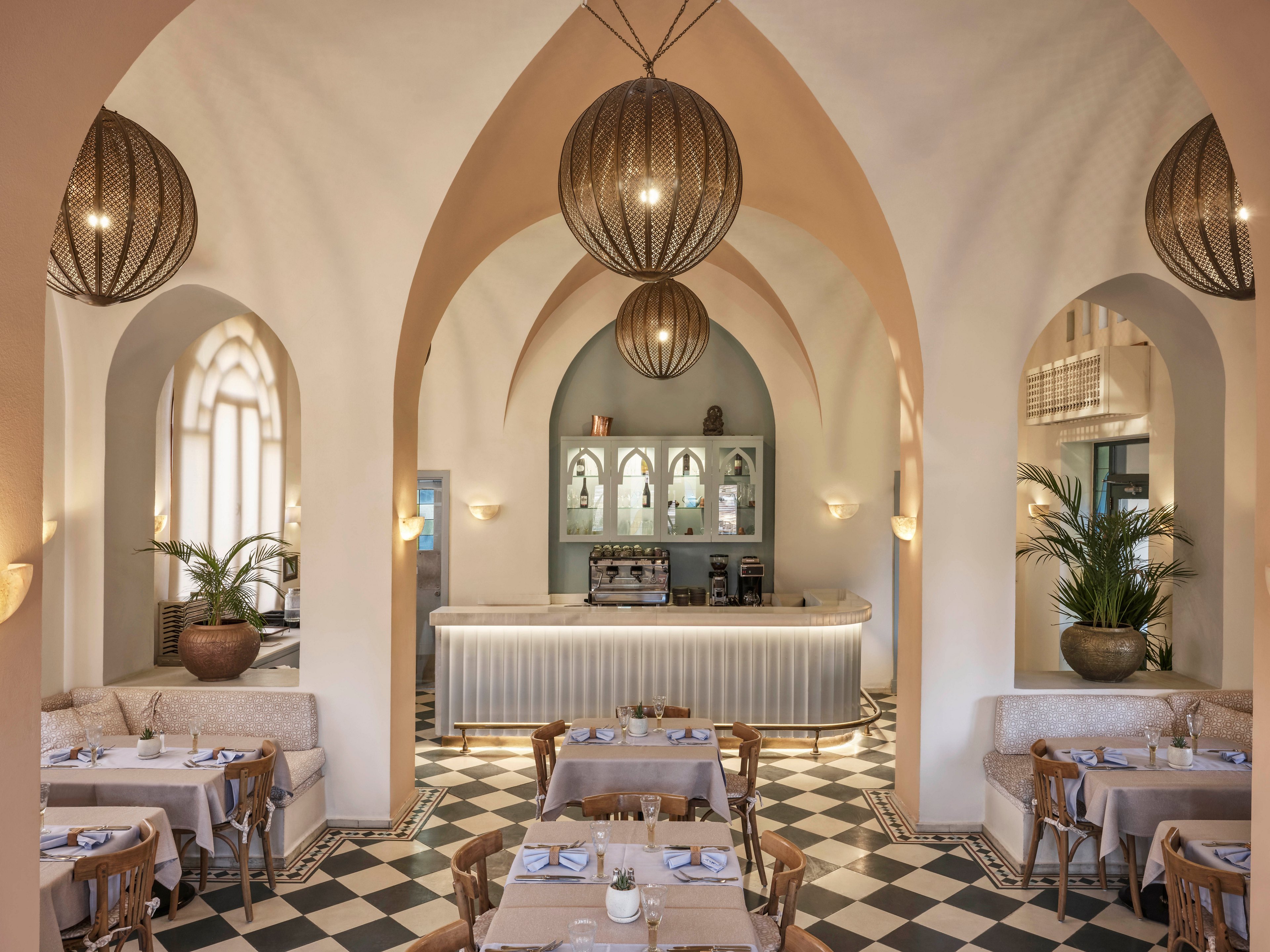 A stylish restaurant interior features high arched ceilings and large pendant lights, creating a warm and inviting atmosphere. The space is decorated with neutral tones, checkered tile flooring, and lush potted plants. Tables are set for dining, and a central bar area stands at the back, enhancing the sophisticated Mediterranean ambiance.
