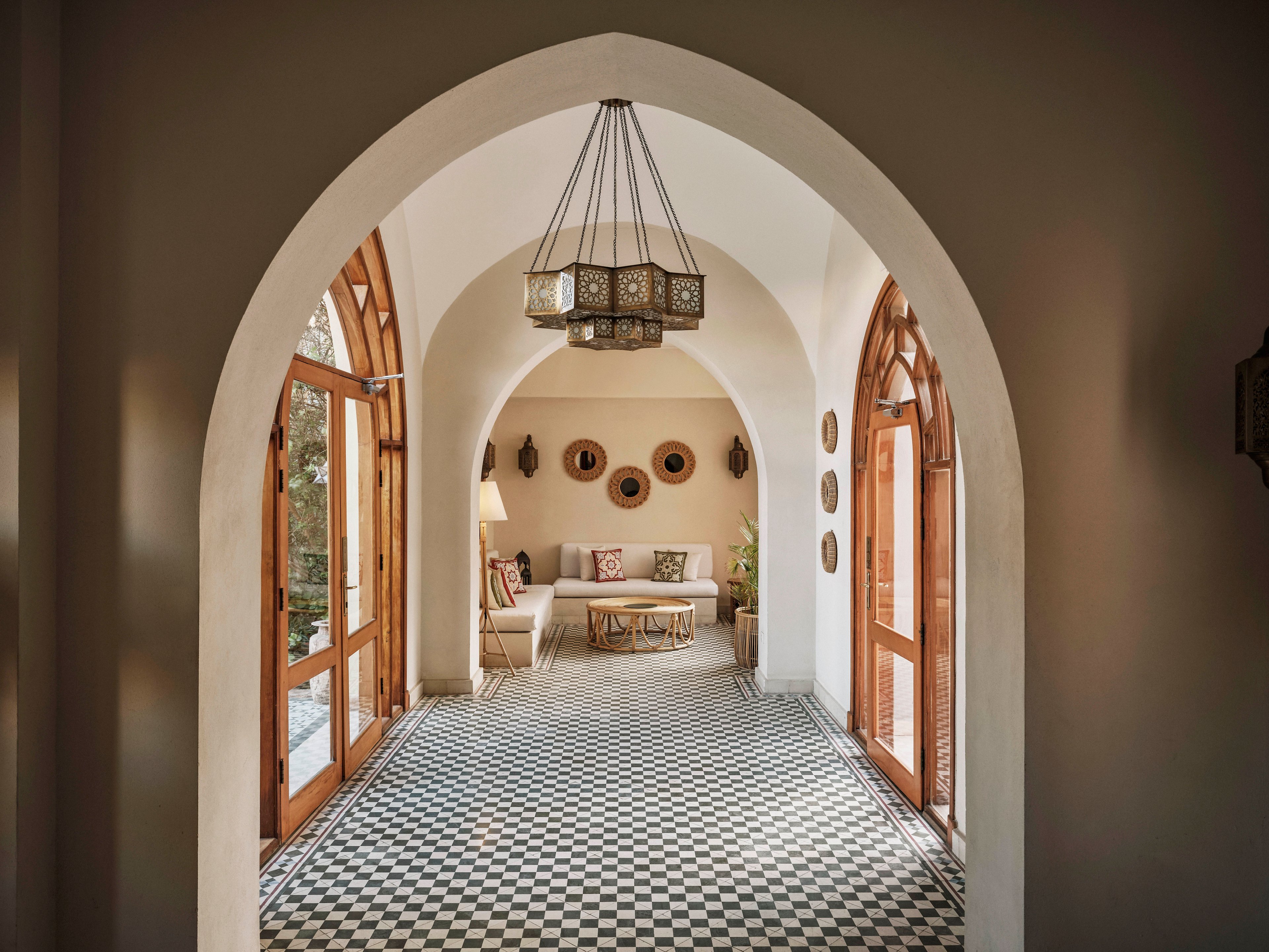 A bright hallway features a distinctive arched entry leading to a cozy sitting area. The space is adorned with patterned black and white floor tiles, wooden framed glass doors, and decorative wall baskets. Natural light fills the room, highlighting the neutral palette and traditional hanging lantern. The setting evokes a tranquil, inviting mood with a blend of modern and Moroccan influences.