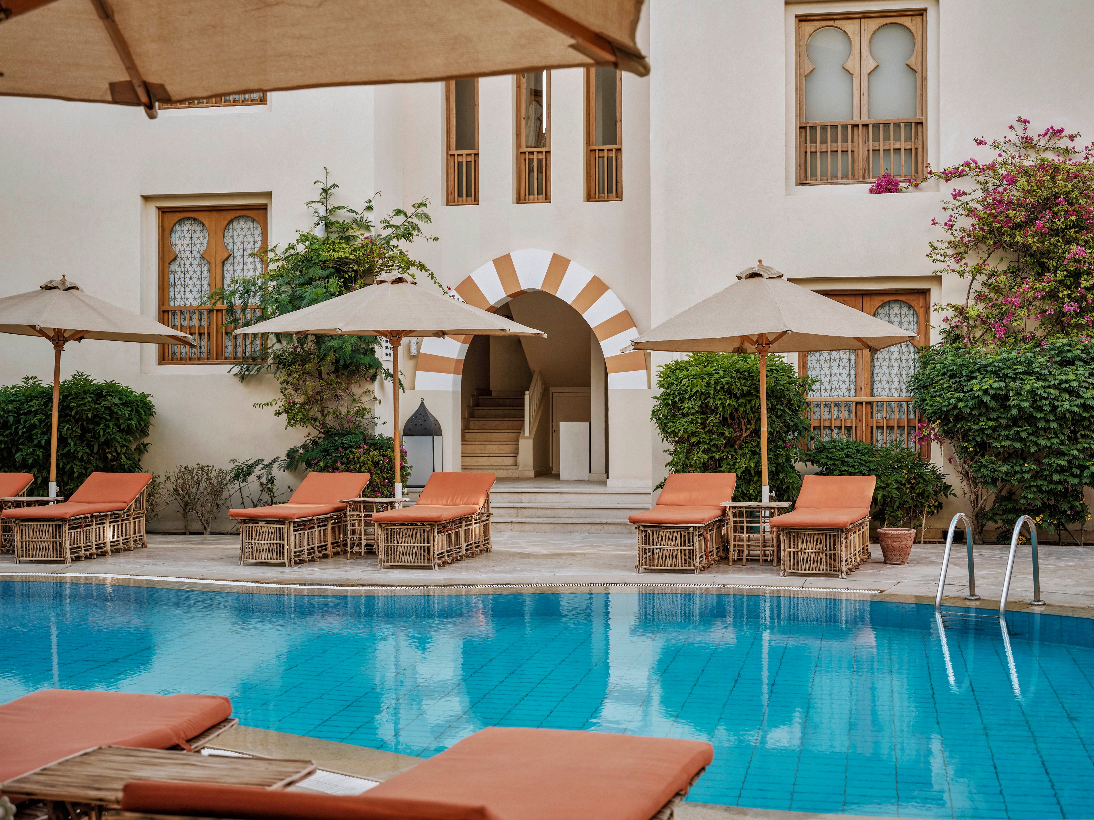 A serene hotel courtyard features a clear blue swimming pool surrounded by wicker sun loungers with orange cushions. Large beige umbrellas provide shade, and lush greenery adds to the tranquil atmosphere. The building’s arched windows and doors showcase elegant architectural details, while the pool ladder invites relaxation. No visible text or numeric values are present in the image.