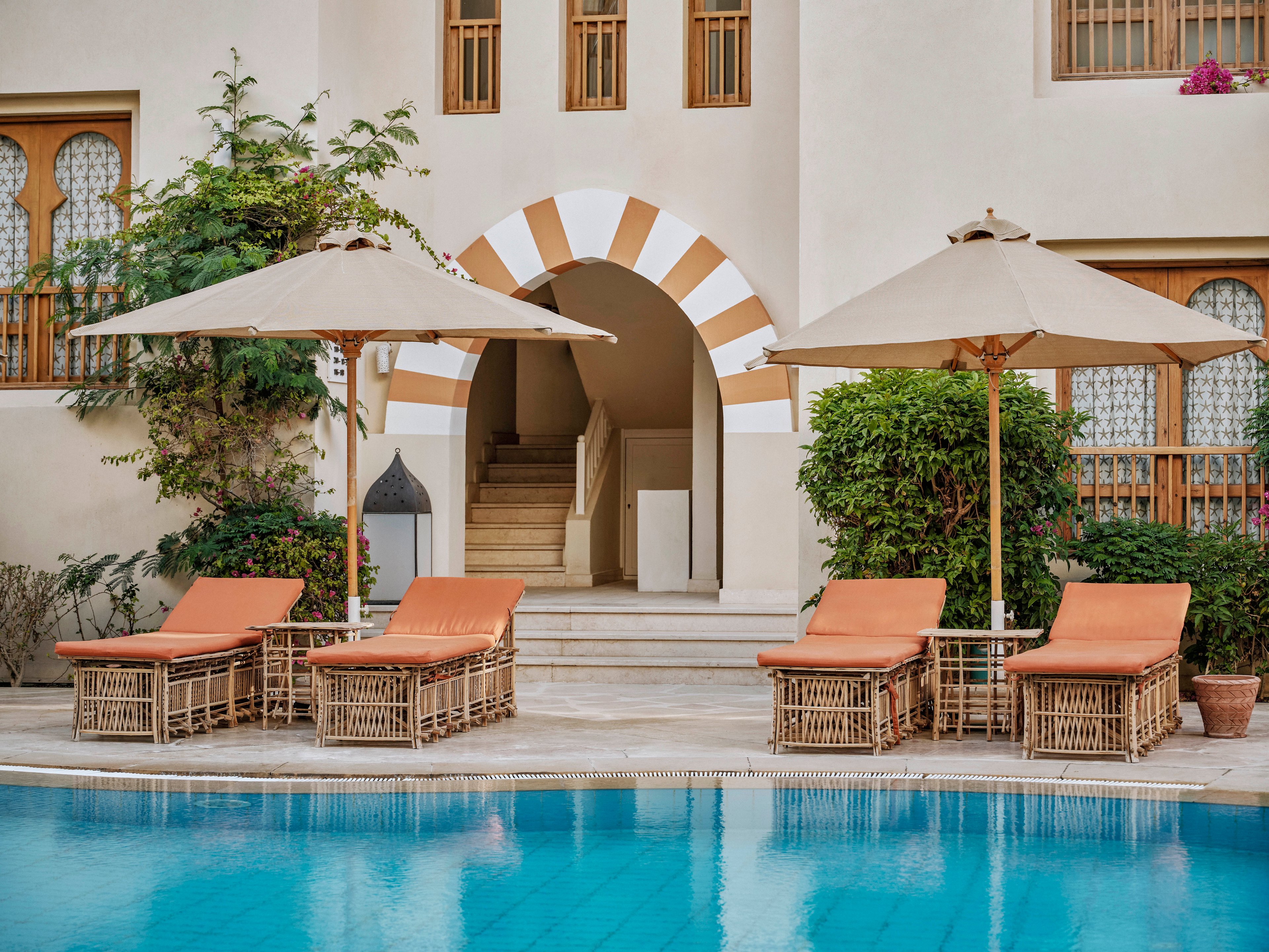 Four wicker loungers with orange cushions are arranged beside a clear blue swimming pool, shaded by two large beige umbrellas. The setting is a tranquil resort courtyard with arched architecture and lush greenery. The scene is calm and inviting, with warm neutral tones and Mediterranean-inspired design elements. No people are present, emphasizing relaxation and leisure.