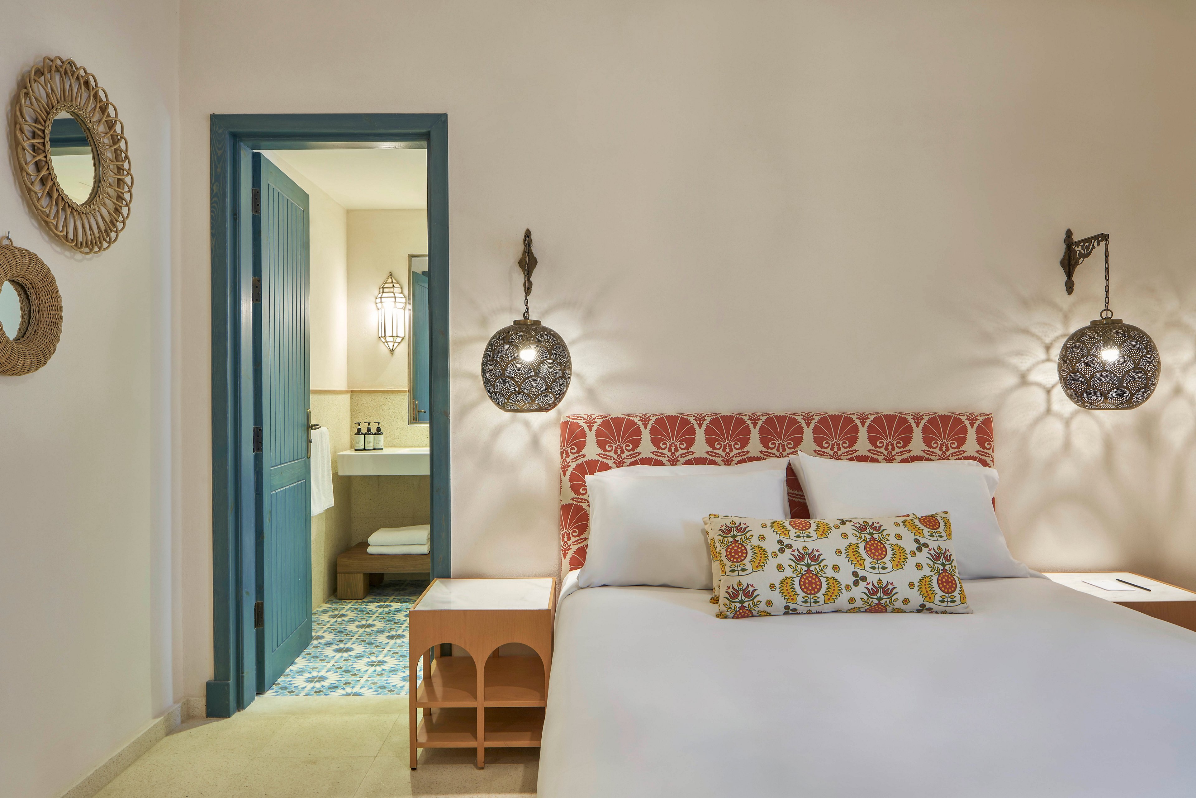 A neatly arranged hotel bedroom features a double bed with a red patterned headboard and a decorative pillow. The room is softly lit with wall-mounted lamps and includes a small wooden side table. The bathroom, visible through a blue door, displays patterned floor tiles and a well-lit vanity area. Decorative mirrors and neutral wall tones create a welcoming, modern atmosphere.