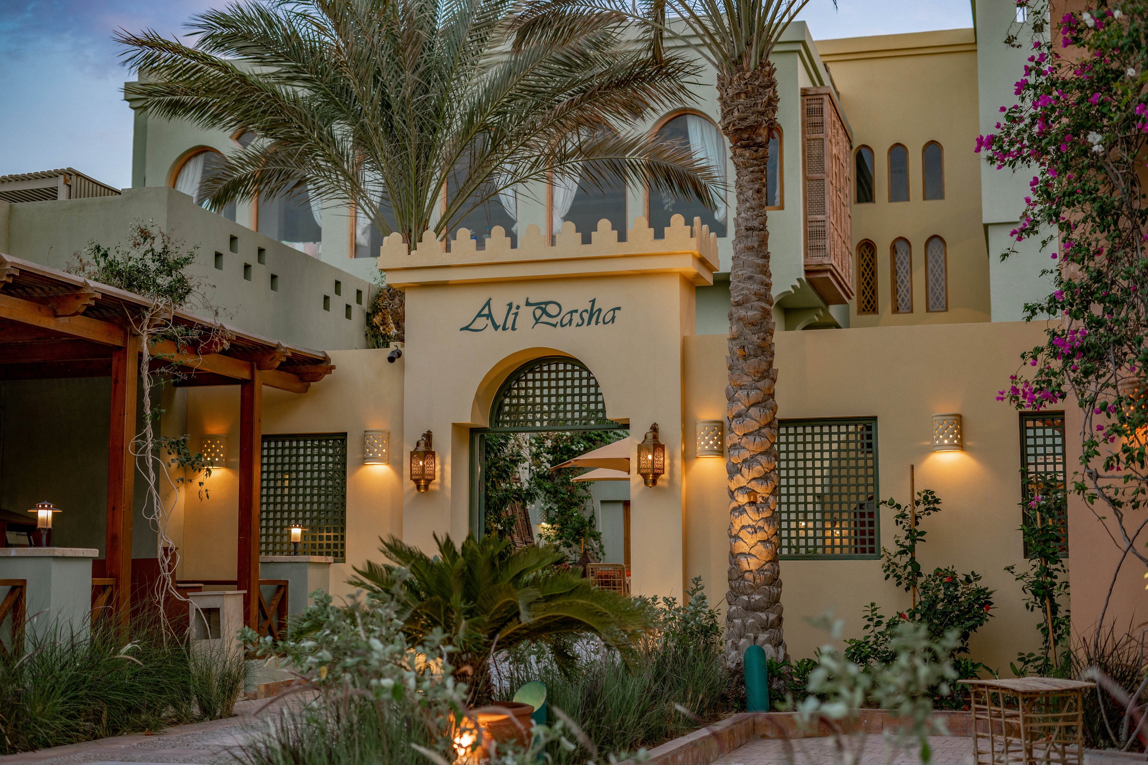 The image shows the entrance of a building with the name 'Ali Pasha' clearly visible above the archway. The setting features lush landscaping, palm trees, and warm ambient lighting, suggesting a tranquil and inviting atmosphere. Architectural details include arched windows, latticework, and traditional Middle Eastern design elements. The scene is captured at dusk, enhancing the serene mood with soft, natural colors.