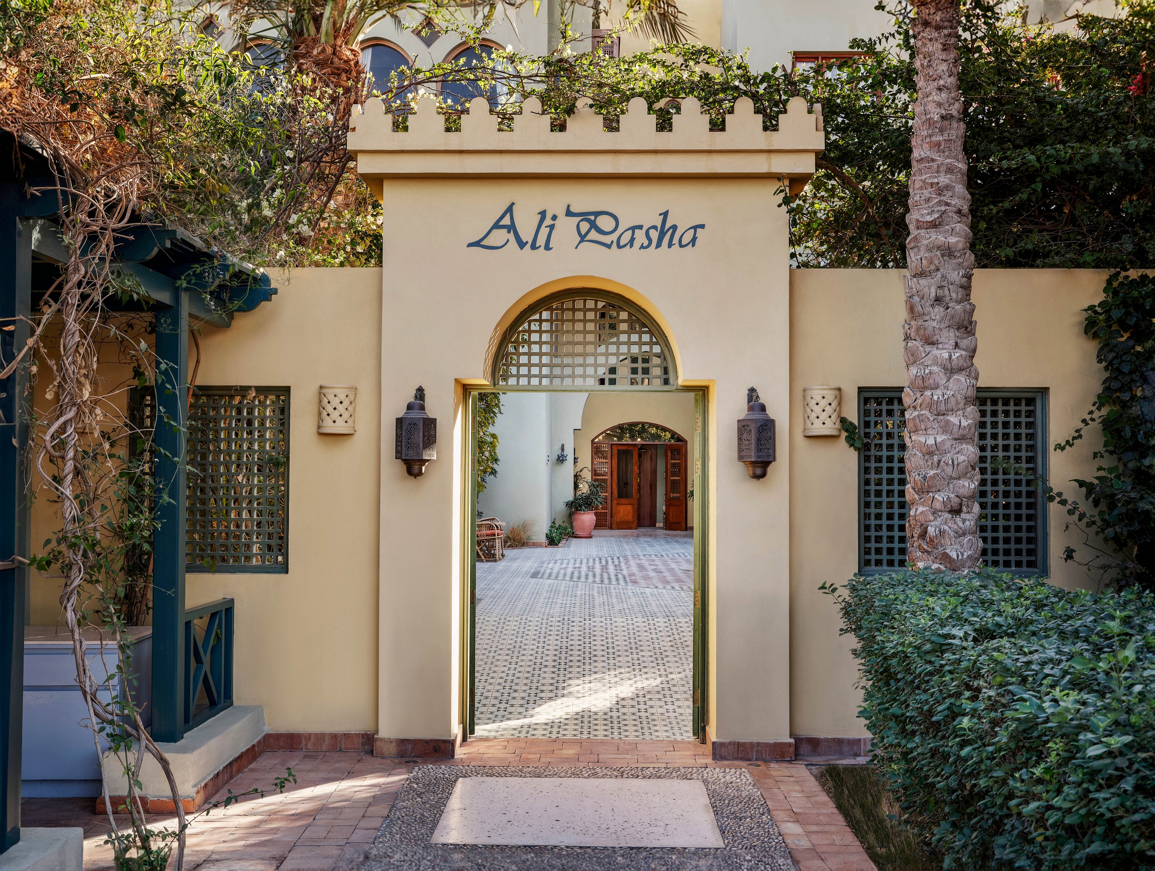 The image shows the entrance to a building named 'Ali Pasha,' featuring a cream-colored archway with decorative battlements. The setting includes lush greenery, palm trees, and a tiled pathway leading to a wooden door in the background. The scene is well-lit with natural daylight, and the overall mood is inviting and tranquil.
