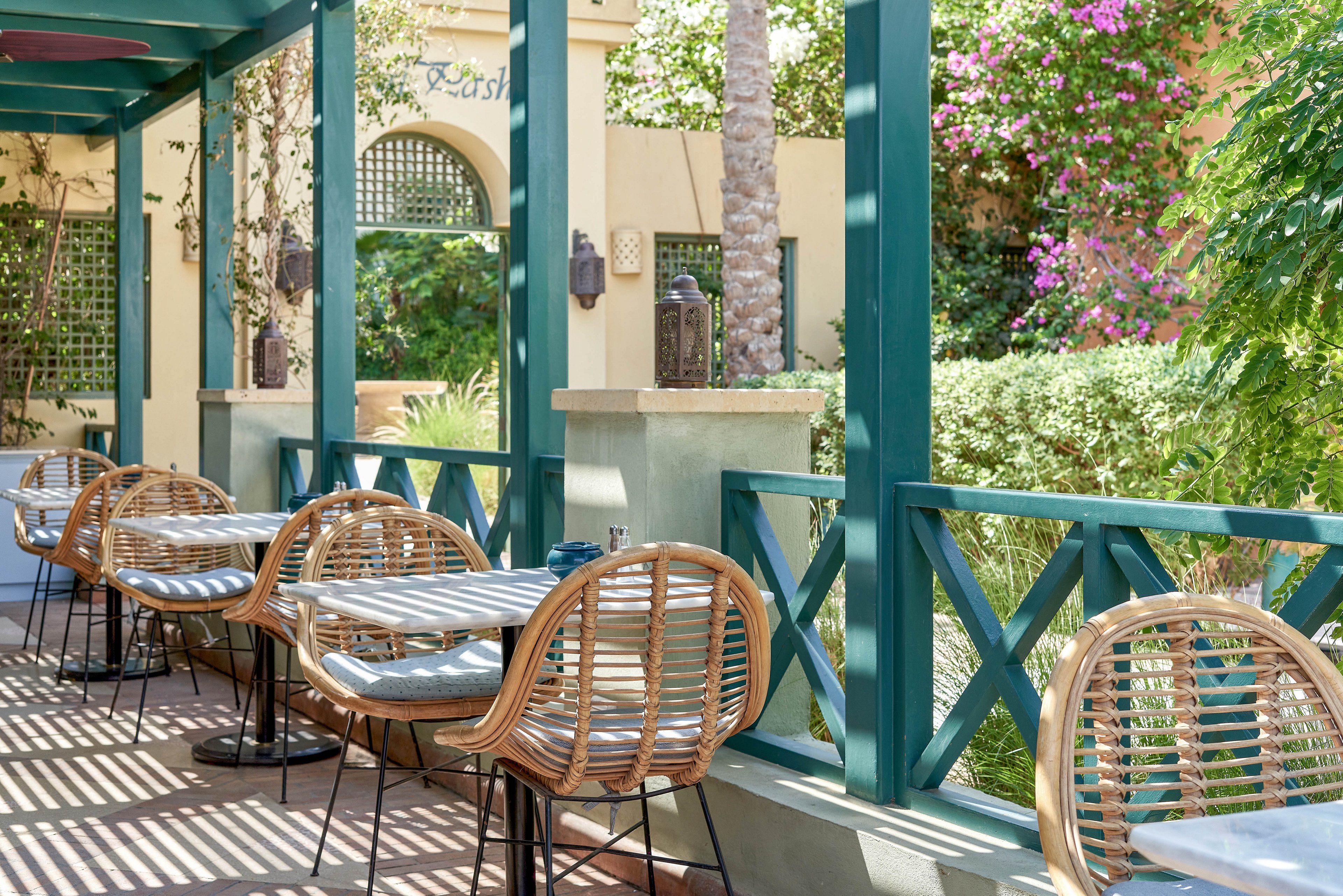 A sunlit outdoor café terrace features several rattan chairs and tables arranged neatly along a green wooden railing. The setting is surrounded by lush greenery, flowering plants, and palm trees, creating a relaxed and inviting atmosphere. Shadows from the pergola overhead create striped patterns on the floor, enhancing the tranquil mood. No visible text or numbers are present in the image.
