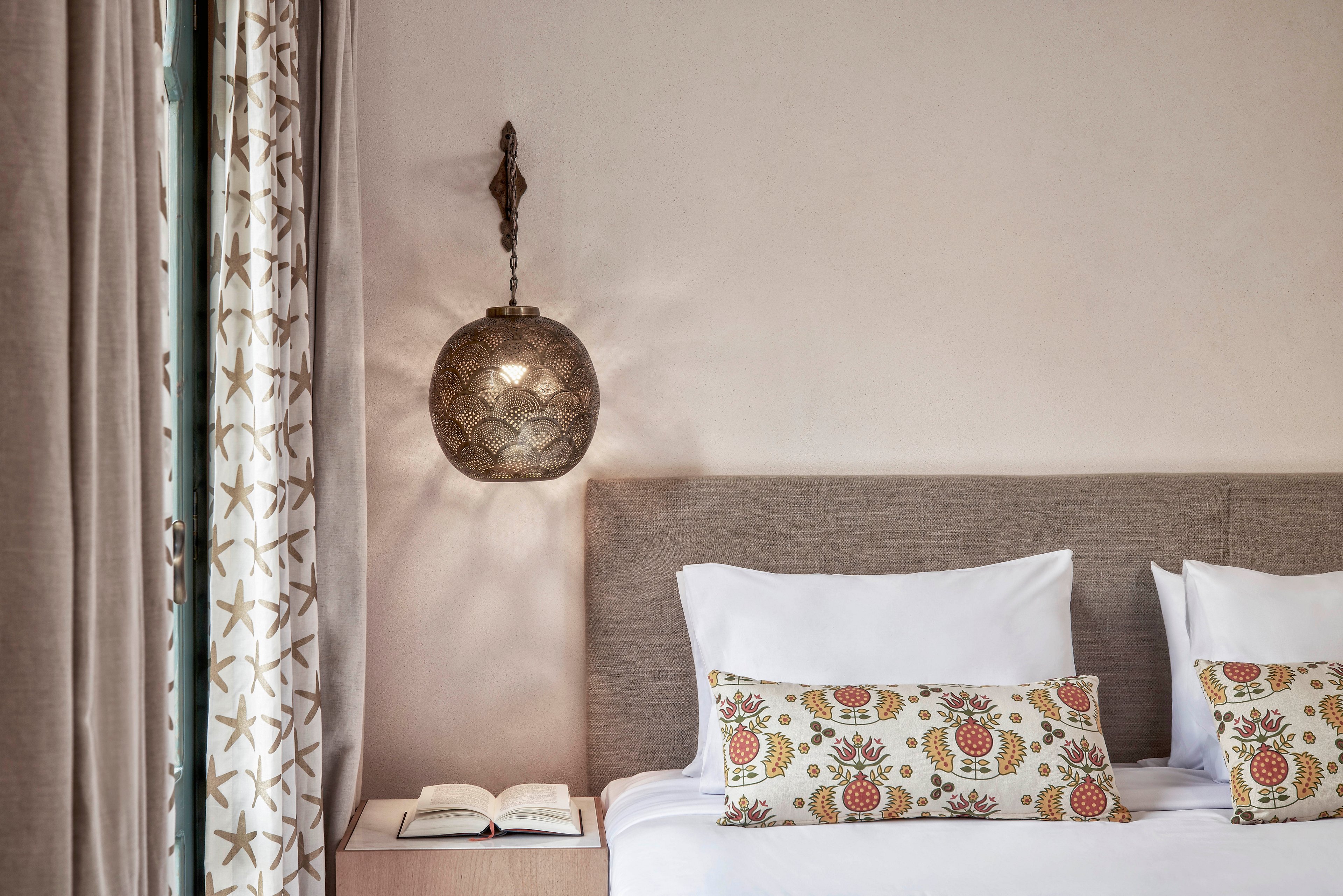 A neatly made bed with white linens and decorative pillows featuring a floral pattern is the main focus. The setting is a softly lit, modern bedroom with neutral tones and a hanging metallic lamp above a bedside table. An open book rests on the table, and patterned curtains add visual interest to the scene.