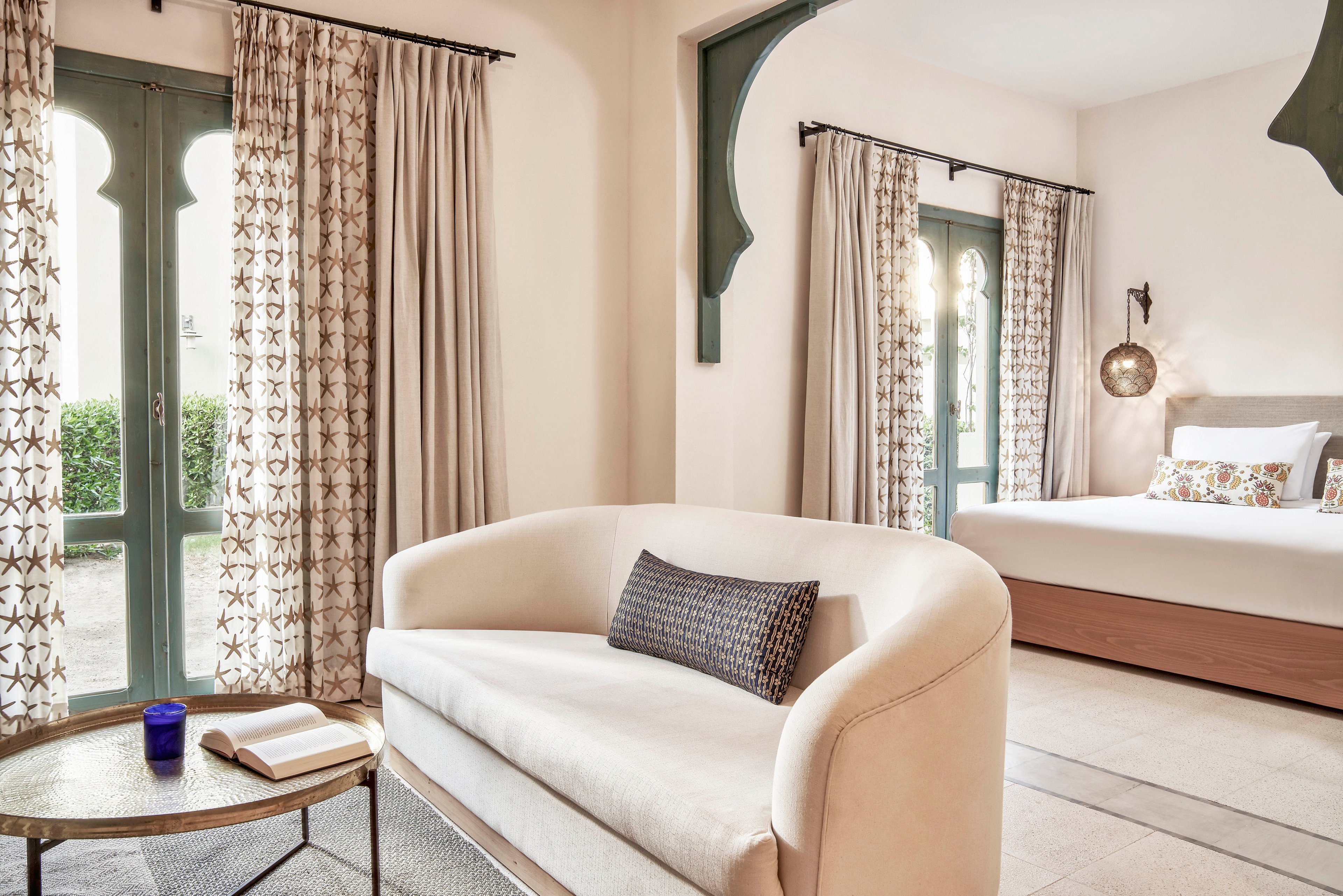 A spacious, sunlit hotel suite features a plush cream sofa and a cozy bed with patterned pillows. Large arched windows with decorative curtains allow natural light to fill the room, highlighting neutral tones and soft textures. A small round table holds a blue cup and an open book, adding a touch of relaxation to the inviting setting. The overall mood is calm and sophisticated, with a blend of contemporary and classic design elements.