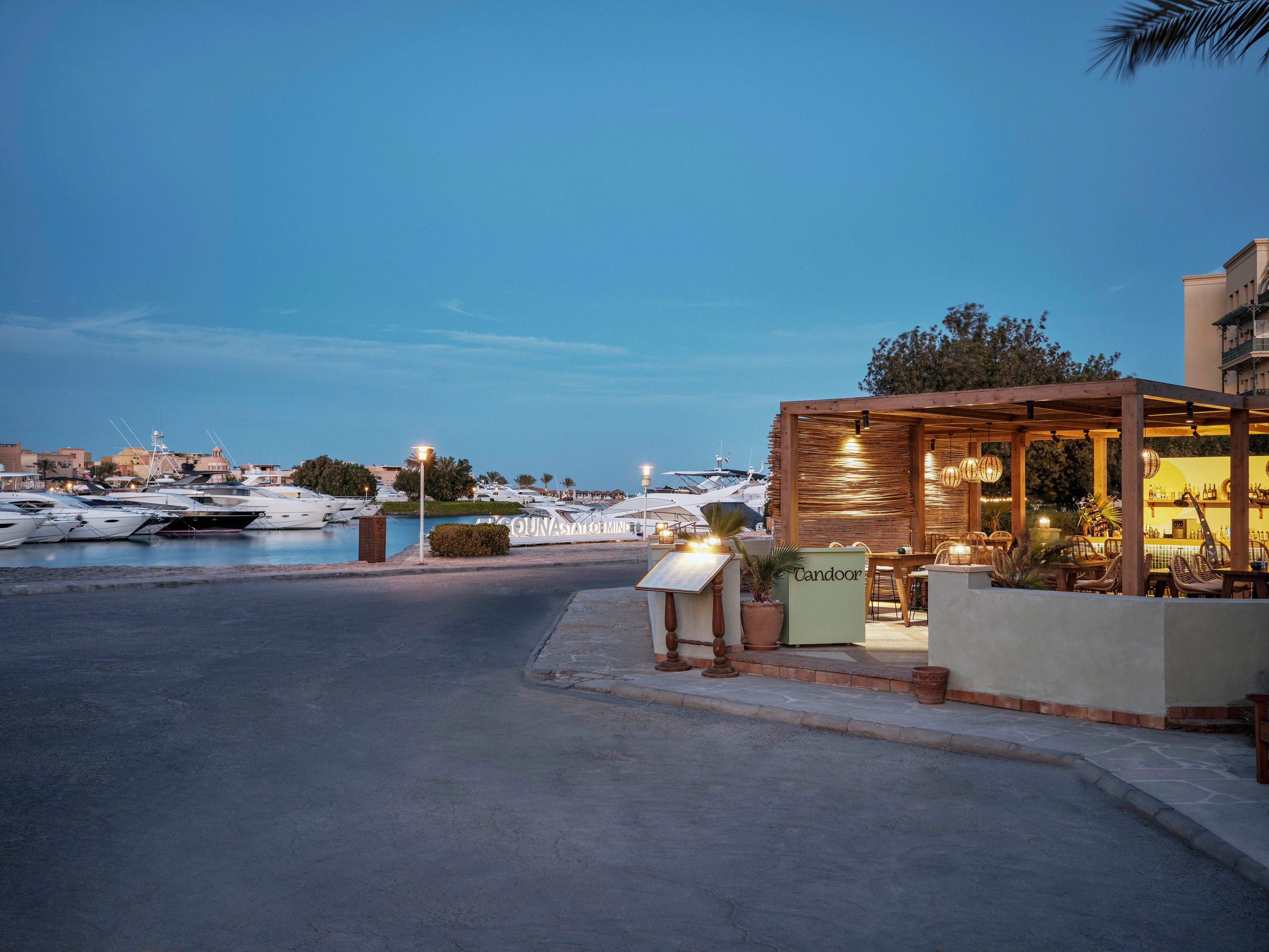 A cozy outdoor restaurant sits beside a marina filled with yachts during twilight. The setting features warm lighting, wooden decor, and a tranquil waterfront view. The restaurant sign reads 'Laudato', and the scene is calm with a clear blue evening sky. No people are visible, emphasizing the peaceful ambiance and inviting atmosphere.