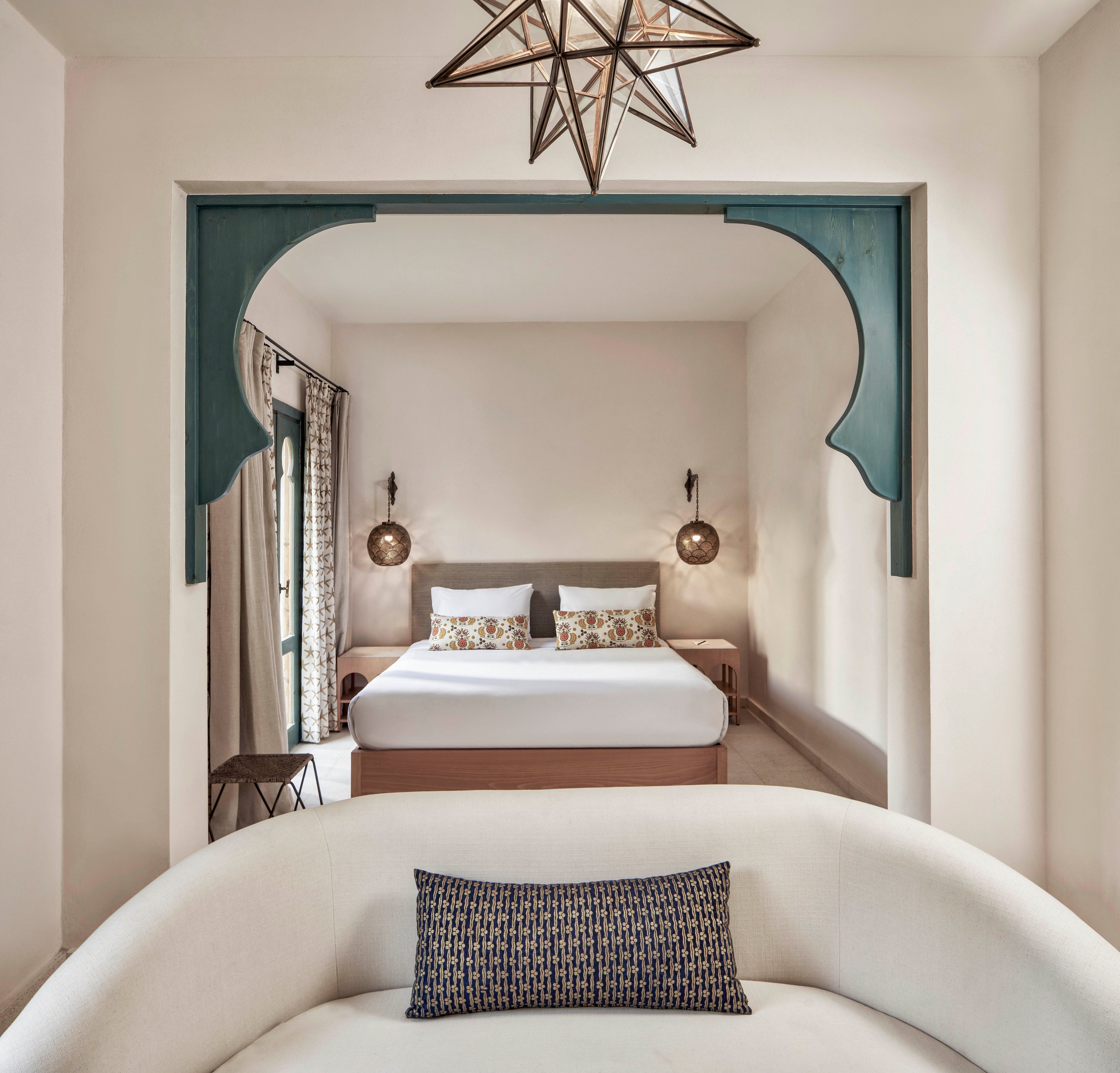 A contemporary bedroom features a neatly made bed with patterned pillows, flanked by two hanging pendant lights. The space is framed by an arched entryway with teal accents and a geometric star-shaped ceiling light. A curved white sofa with a decorative cushion sits in the foreground, while natural light filters through a window with patterned curtains.