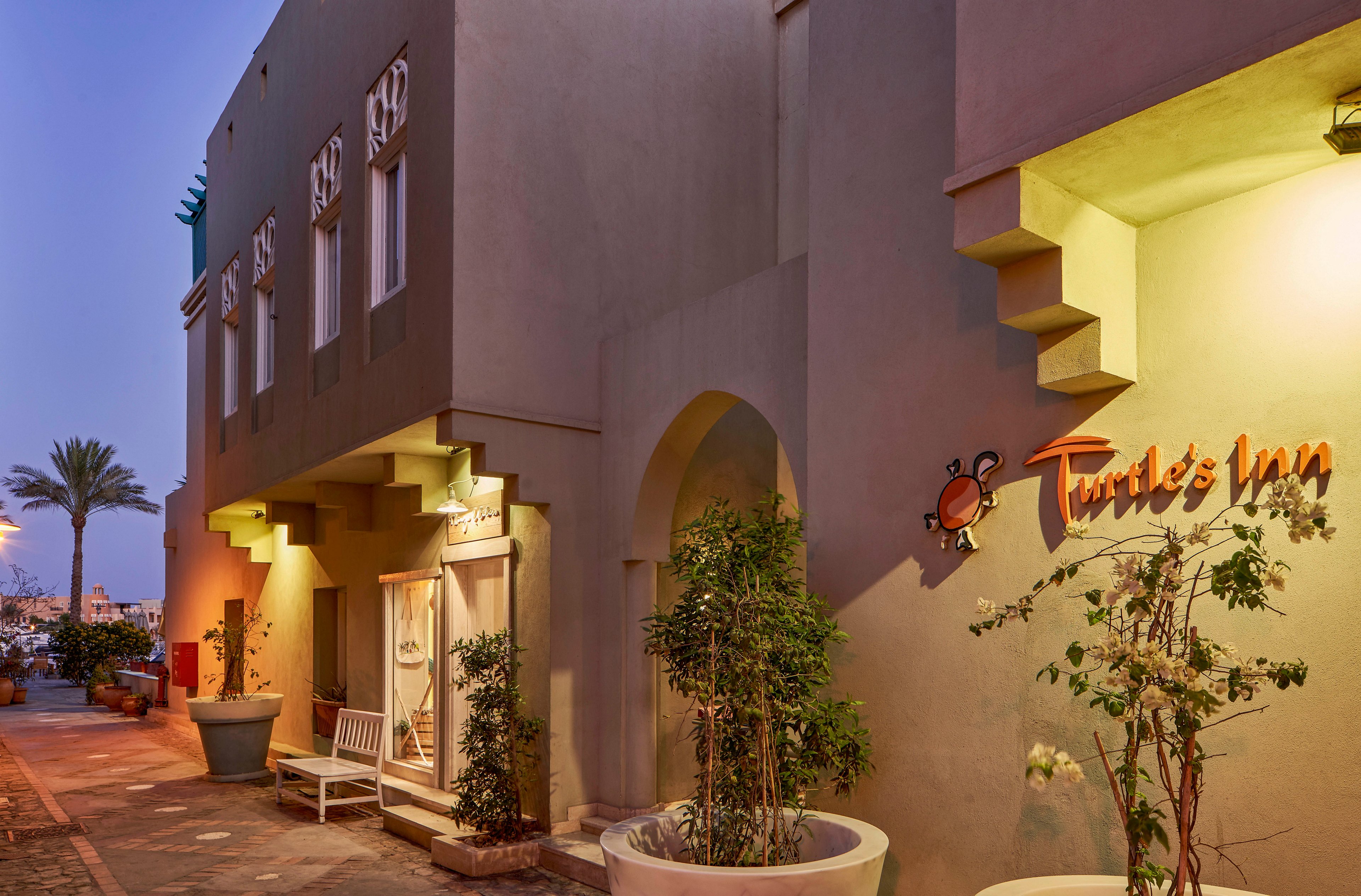 The image shows the exterior of Turtle's Inn, a building with a modern architectural style and soft, warm lighting at dusk. The inn's name and turtle graphic are clearly visible on the wall. Potted plants and benches line the sidewalk, creating a welcoming atmosphere. The sky is a gradient of blue and purple, and a palm tree is visible in the background.
