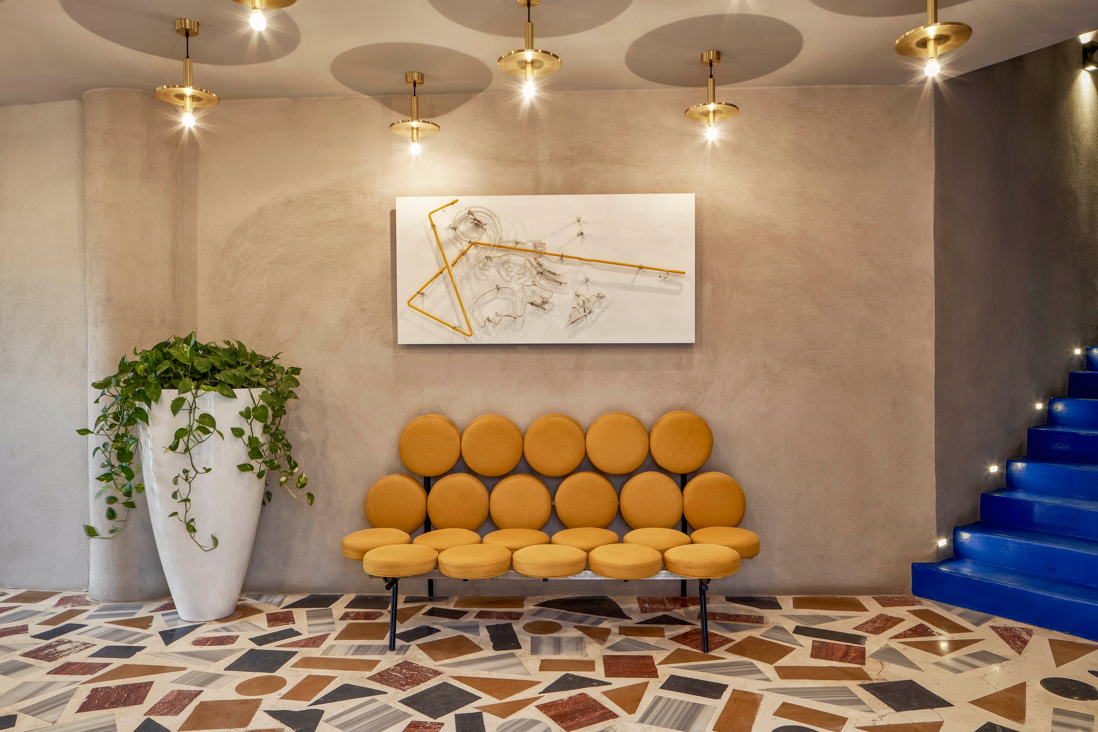 A contemporary lobby features a unique circular-cushion bench in mustard yellow, set against a neutral wall. Above the bench hangs an abstract artwork with gold accents. The space includes a large white planter with trailing greenery and a striking blue staircase with illuminated steps. The floor is decorated with a colorful geometric tile pattern, and round ceiling lights add a warm ambiance.