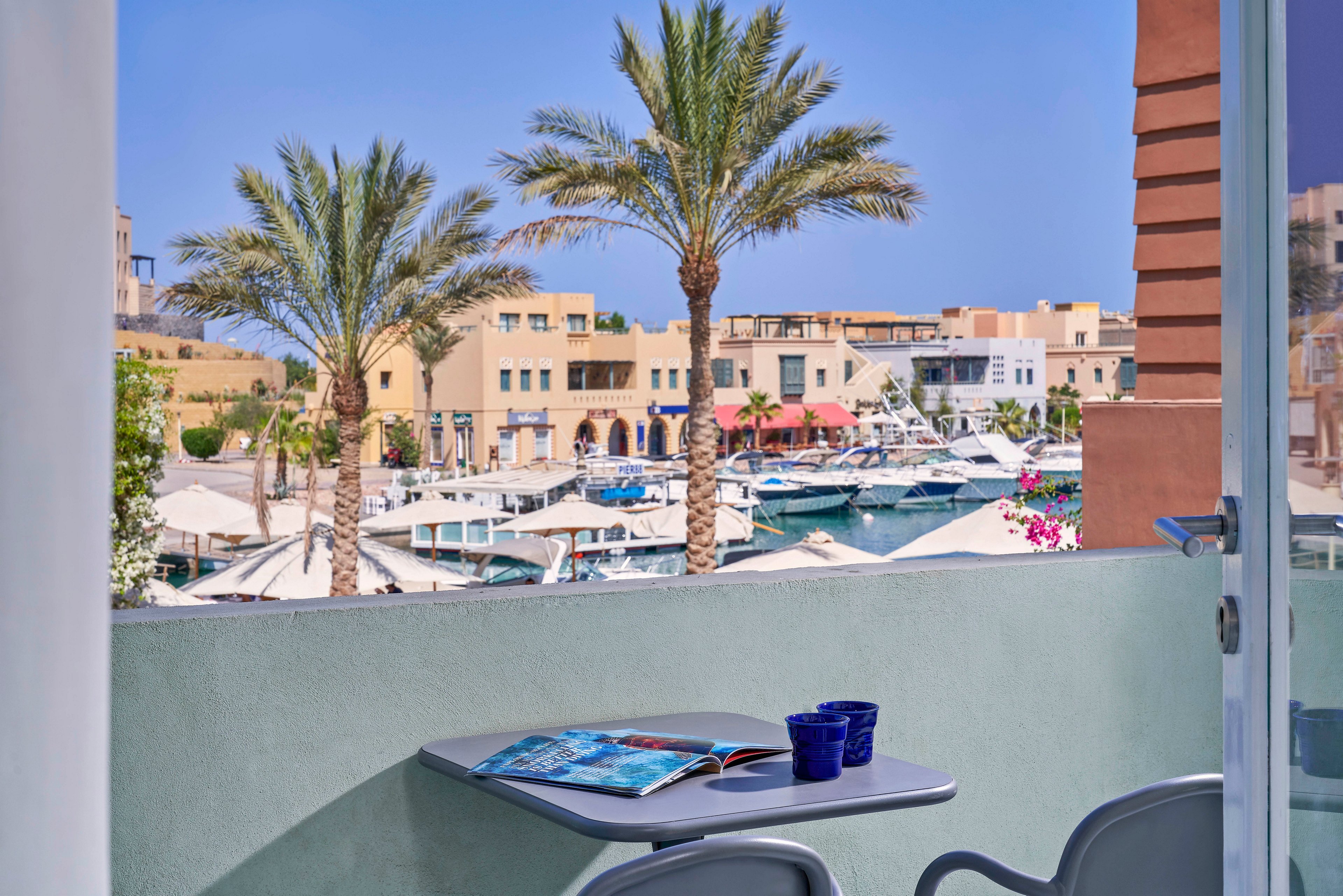 A balcony scene overlooks a vibrant marina filled with boats and yachts, framed by tall palm trees and Mediterranean-style buildings. On the balcony table, two blue cups and an open magazine suggest a relaxing moment. The setting is bright and sunny, evoking a leisurely, vacation-like atmosphere.