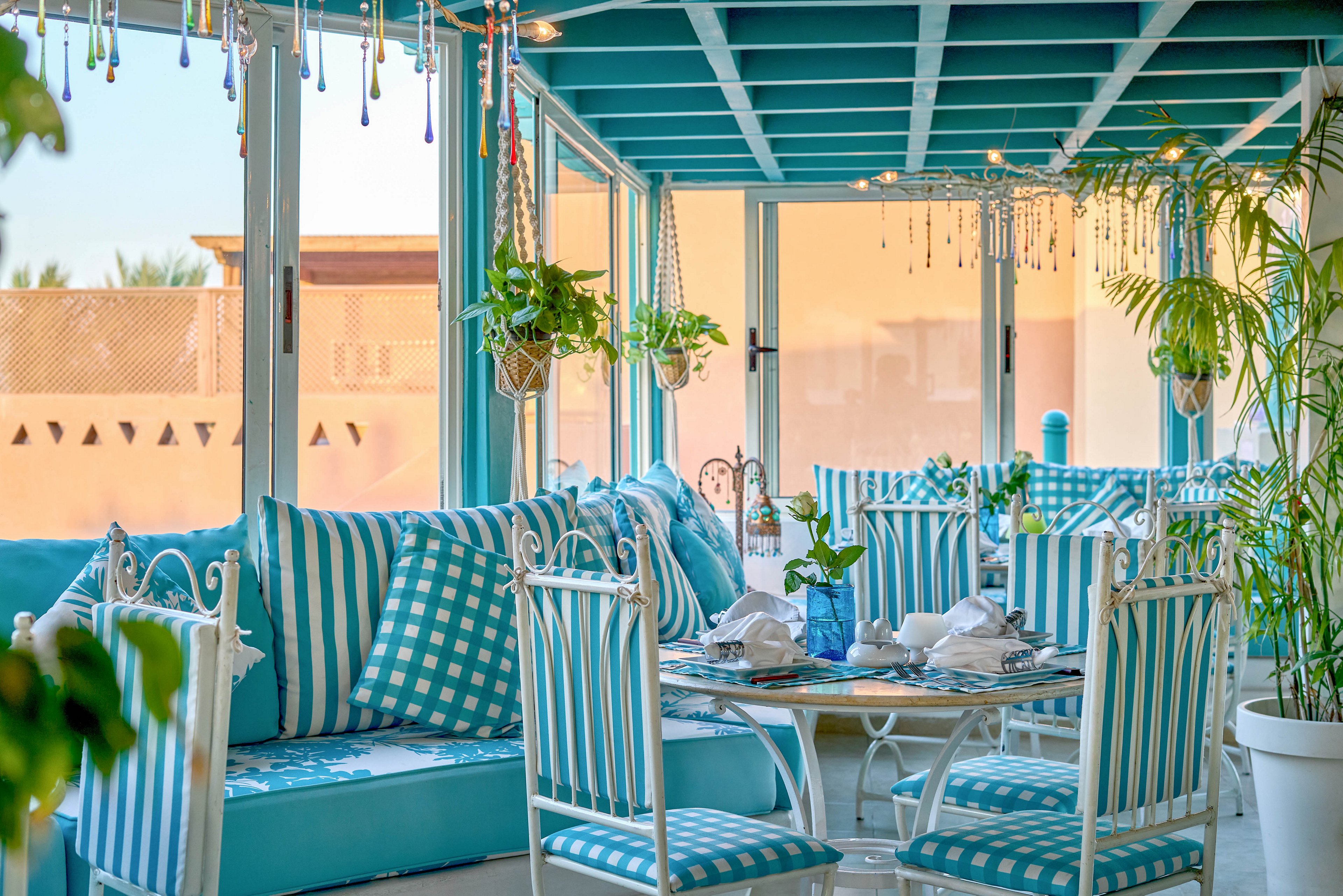A sunlit cafe interior features turquoise and white striped chairs and cushions, creating a fresh and inviting atmosphere. The space is decorated with hanging plants, glass bead accents, and large windows that let in natural light. Tables are set with white napkins and glassware, while the overall palette is dominated by cool blue and white tones. The setting suggests a relaxed, coastal-inspired dining experience.