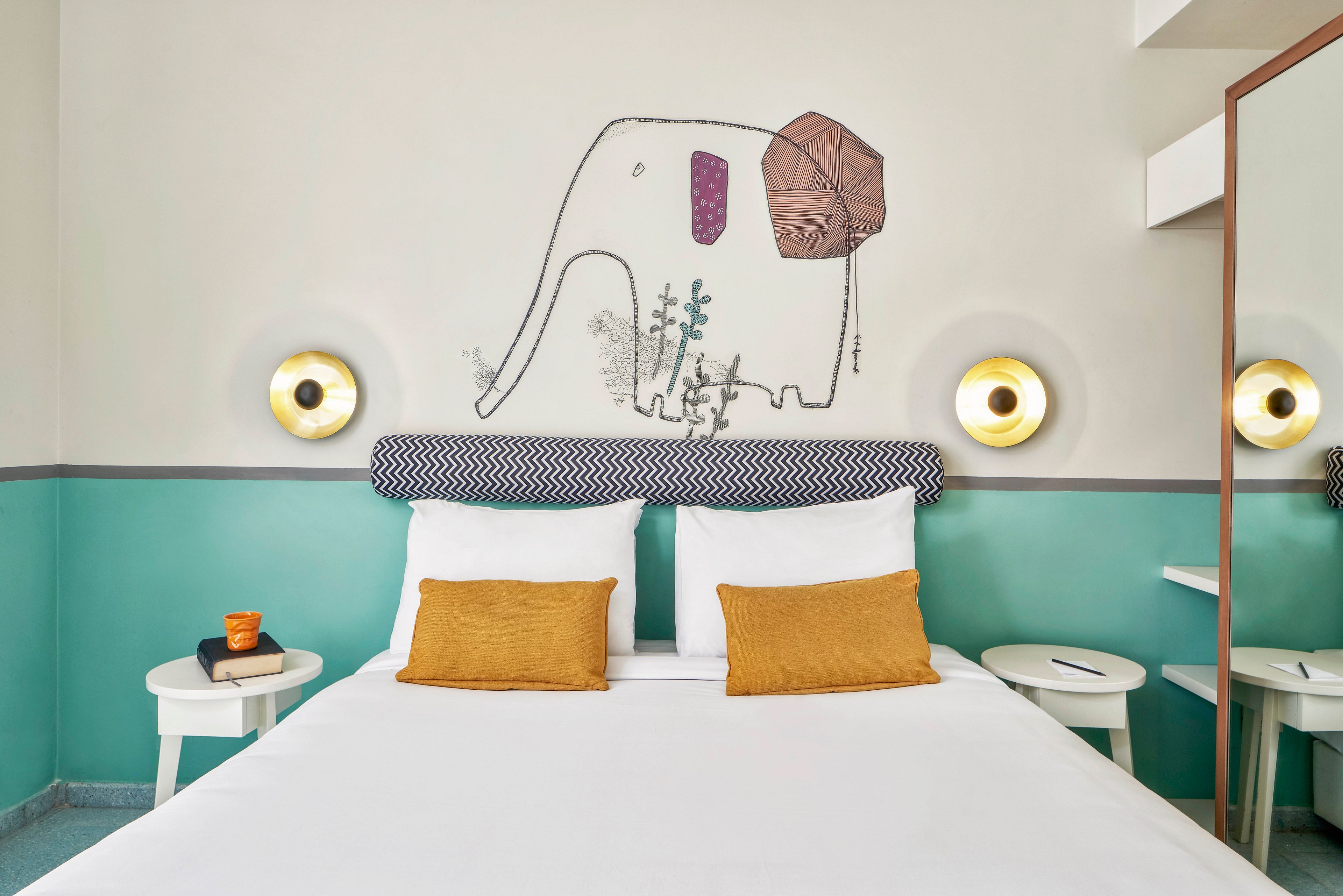 A contemporary hotel room features a double bed with white linens and two mustard yellow accent pillows. The headboard is patterned, and above it is a whimsical elephant illustration on the wall, adding playful character. The room is decorated in soft teal and white tones, with round wall lights and minimalist side tables, creating a fresh and inviting atmosphere.