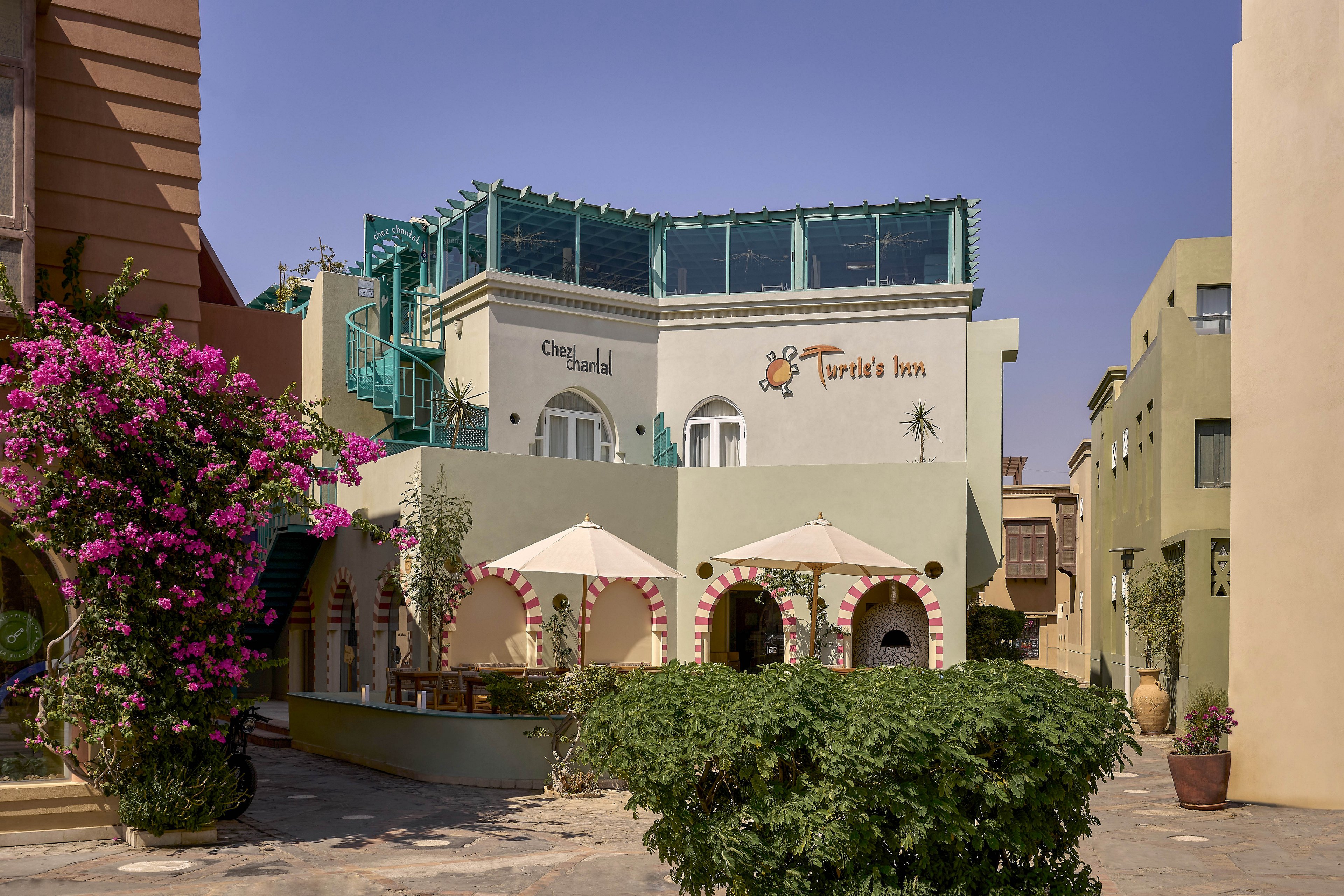 A sunlit courtyard features a pastel-colored building with visible signage for 'Turtle's Inn' and 'Chez Pascal.' The setting includes outdoor seating shaded by large umbrellas, surrounded by flowering plants and Mediterranean-style architecture. The sky is clear, and the overall mood is relaxed and inviting. No people are visible in the scene, and the environment suggests a warm, leisurely atmosphere.